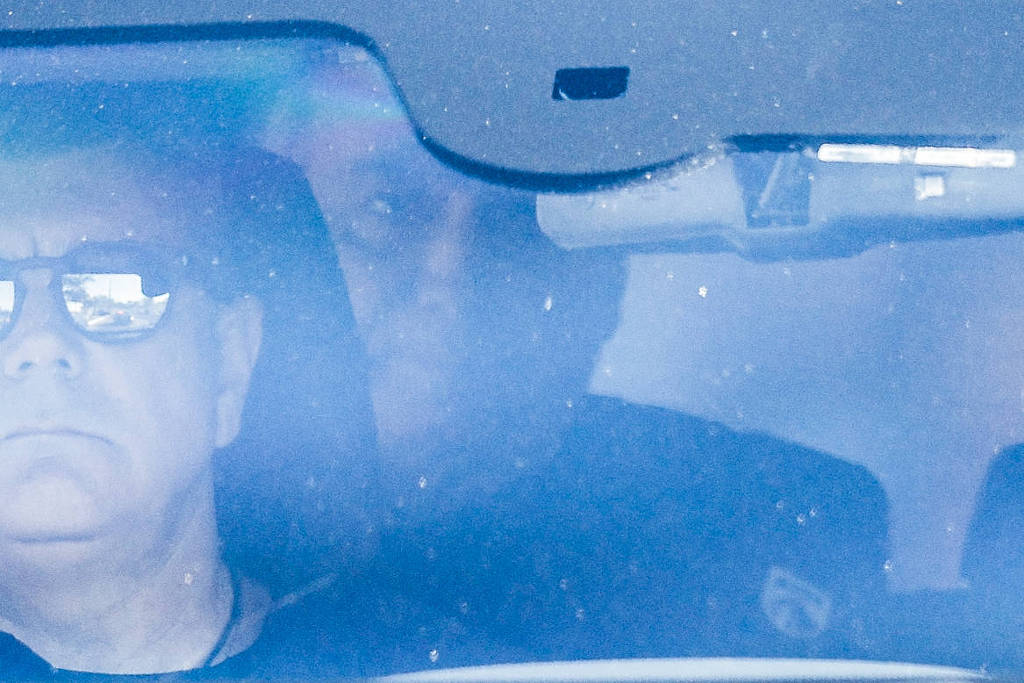 A imagem mostra dois homens dentro de um carro. O homem da frente está usando óculos escuros e parece estar focado na estrada. O segundo homem, que está atrás, é menos visível devido ao reflexo e à iluminação. A imagem tem um aspecto embaçado, possivelmente devido a sujeira no vidro ou reflexos.