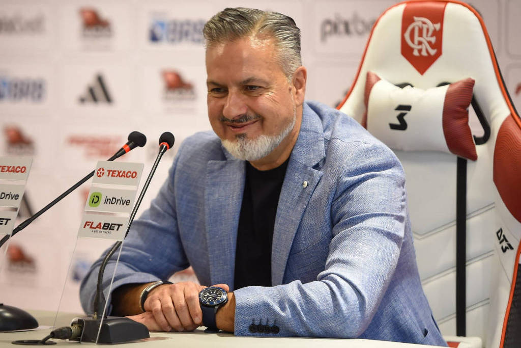 Um homem sorridente está sentado em uma mesa de coletiva de imprensa. Ele usa um blazer claro e uma camiseta escura. Atrás dele, há uma cadeira com o logotipo do Flamengo e um fundo com logotipos de patrocinadores. O homem parece estar em um ambiente de esportes, possivelmente relacionado ao futebol.
