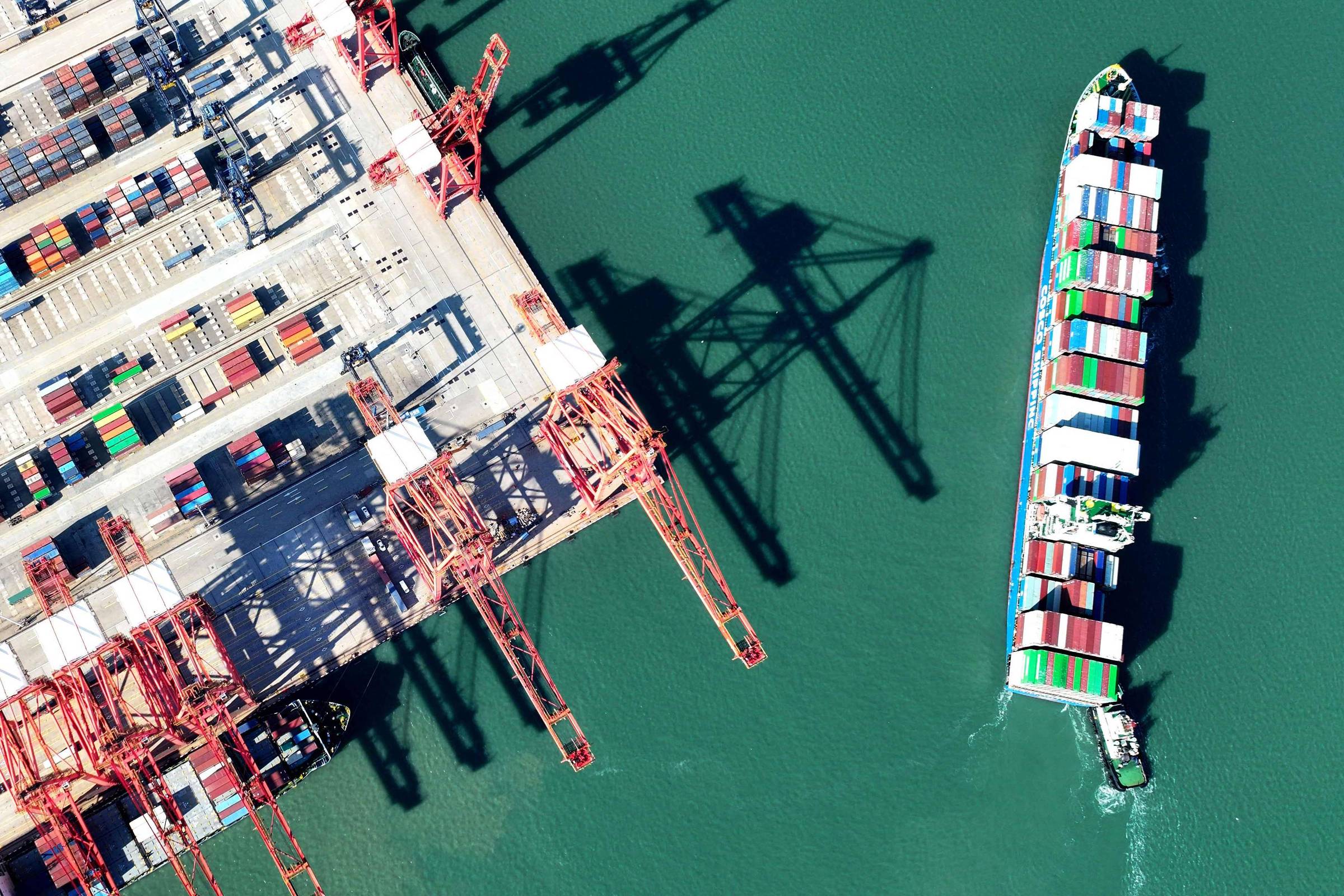 A imagem mostra uma vista aérea de um porto com vários contêineres coloridos empilhados em um cais. À direita, um navio cargueiro está se aproximando do cais, carregando contêineres em sua parte superior. As sombras das gruas e dos contêineres são visíveis na água verde abaixo.