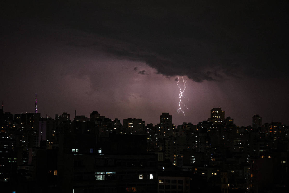 A imagem mostra uma cidade à noite, com muitos prédios iluminados. No céu, nuvens escuras estão presentes, e um raio é visível, iluminando a cena. A atmosfera é de tempestade, com uma iluminação dramática causada pelo relâmpago.