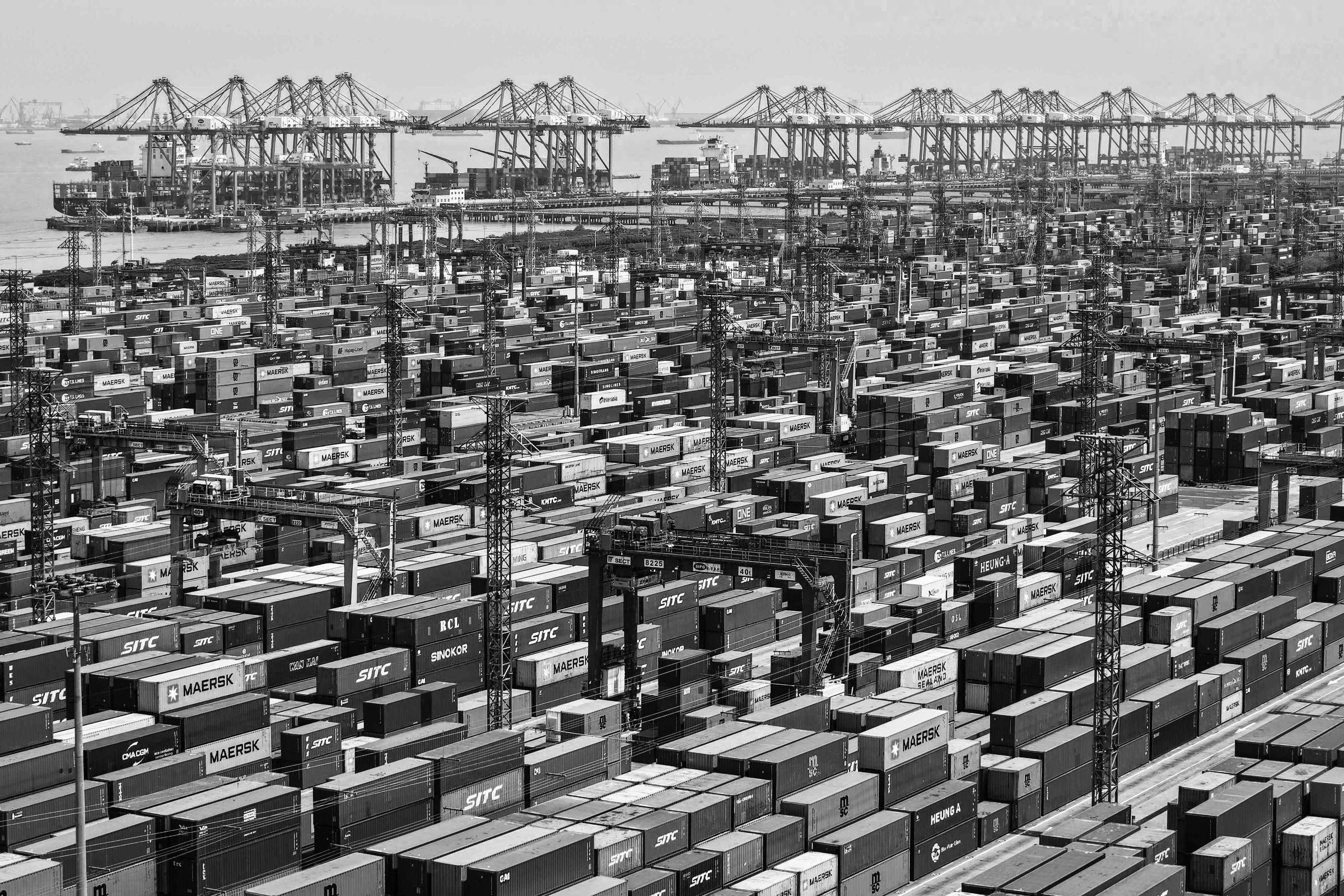 A imagem mostra uma vasta área de um porto de contêineres, repleta de contêineres de diferentes cores e tamanhos organizados em fileiras. Ao fundo, há guindastes vermelhos que se destacam contra o céu nublado, sugerindo atividade portuária intensa. A cena é dominada por uma grande quantidade de contêineres, evidenciando a movimentação de carga.