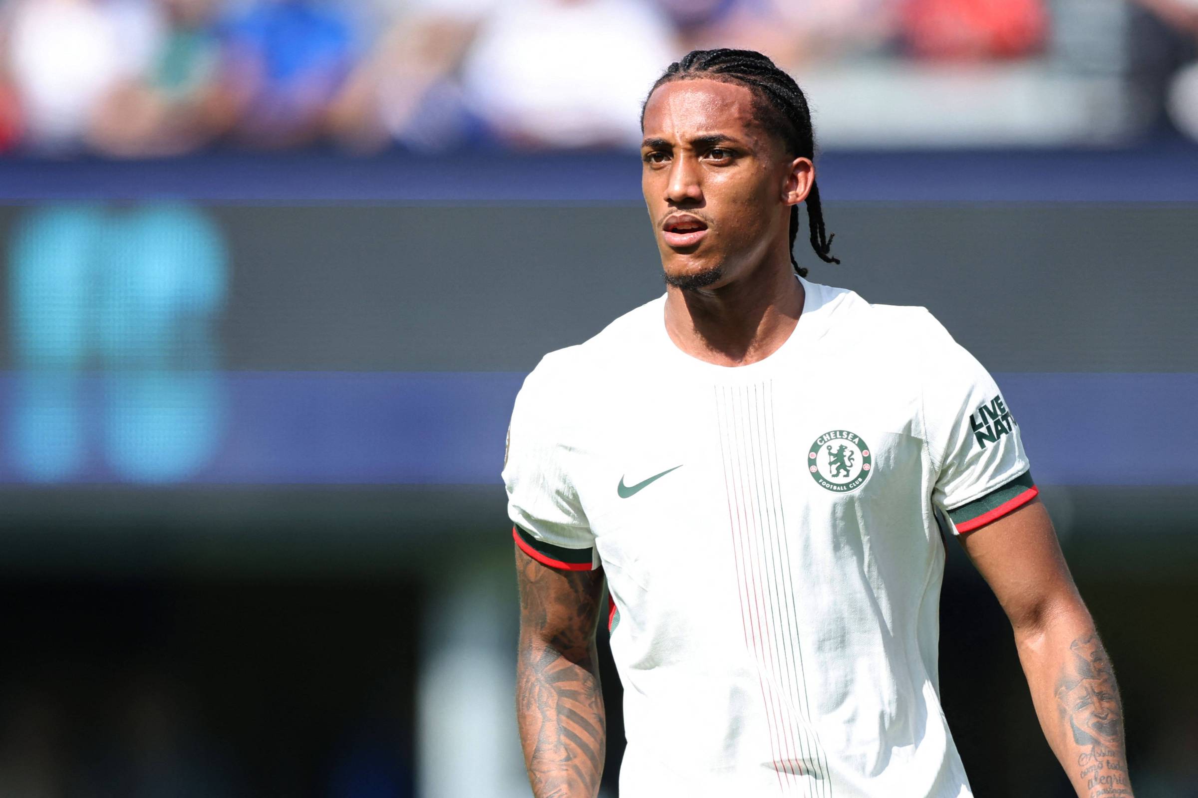 Um jogador de futebol está em campo, vestindo uma camisa branca com detalhes em vermelho e verde. O jogador tem cabelo preso em tranças e está olhando para frente, com uma expressão concentrada. Ao fundo, é possível ver uma multidão e um campo de futebol.