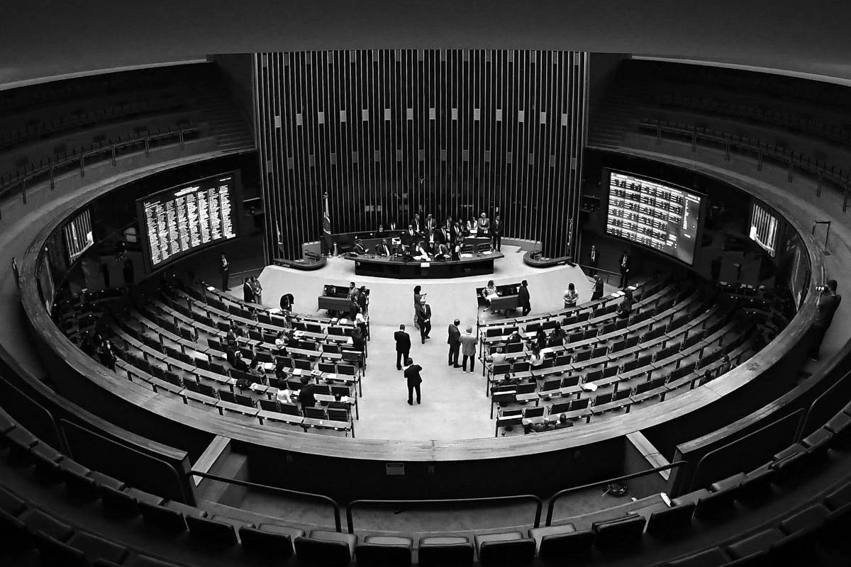 A imagem mostra uma visão panorâmica do plenário do Congresso Nacional do Brasil. O espaço é circular, com assentos dispostos em camadas ao redor de uma área central onde estão algumas pessoas, possivelmente deputados. Ao fundo, há painéis eletrônicos exibindo informações. O teto apresenta um design circular com um padrão em espiral.