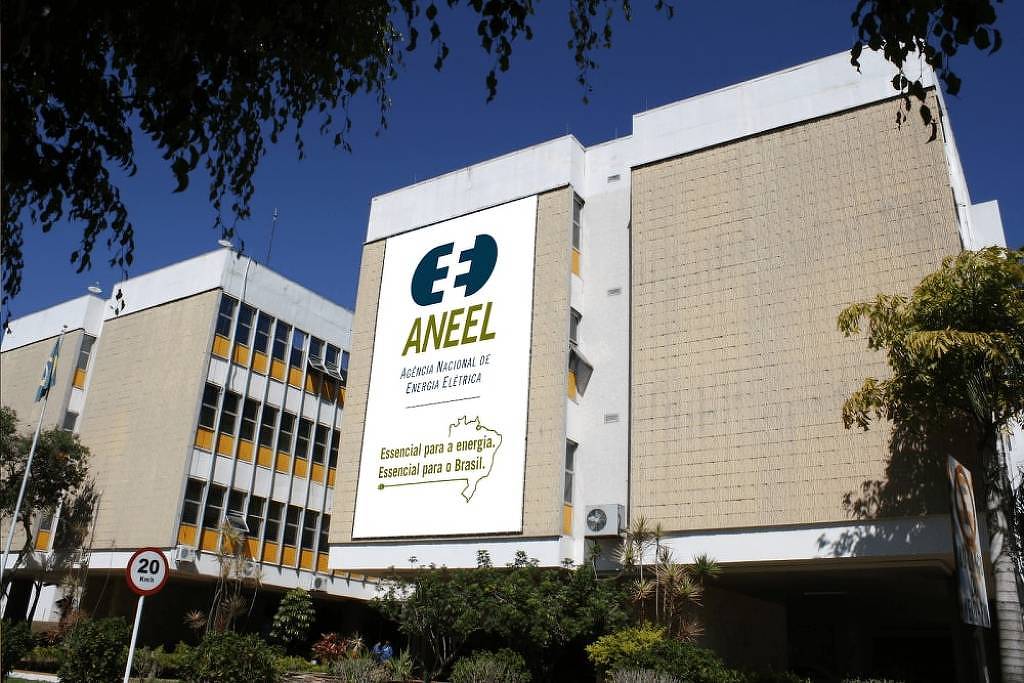 A imagem mostra o edifício da ANEEL (Agência Nacional de Energia Elétrica) em Brasília. A fachada do prédio é de cor clara, com janelas e detalhes em amarelo. Em destaque, há um grande painel com o logotipo da ANEEL e a frase