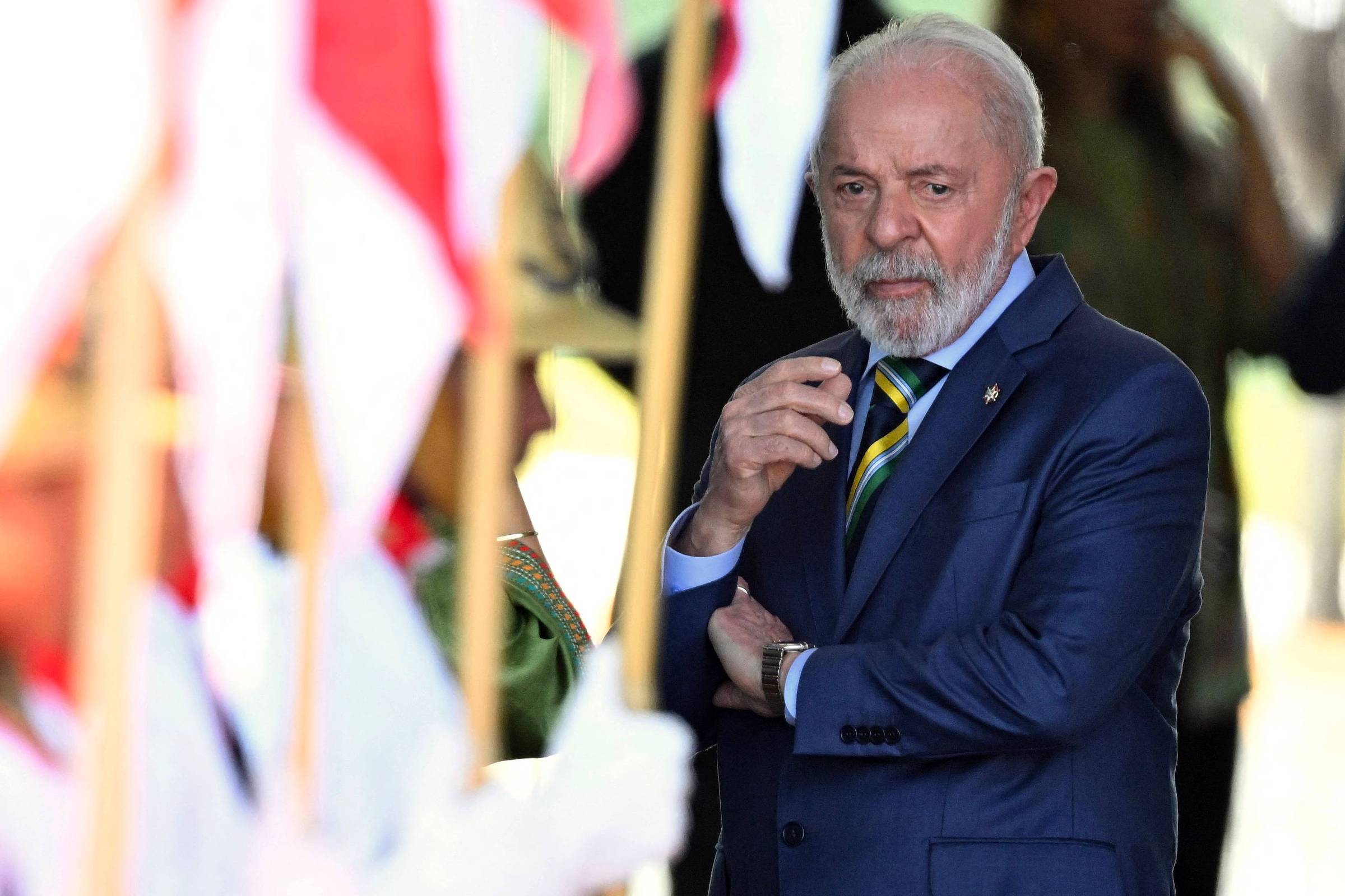 Um homem de terno azul e gravata, com cabelo grisalho e barba, observa atentamente enquanto está em um ambiente de cerimônia. Ao fundo, há pessoas segurando bandeiras, algumas com cores vermelhas e brancas. O homem parece pensativo e está com os braços cruzados.