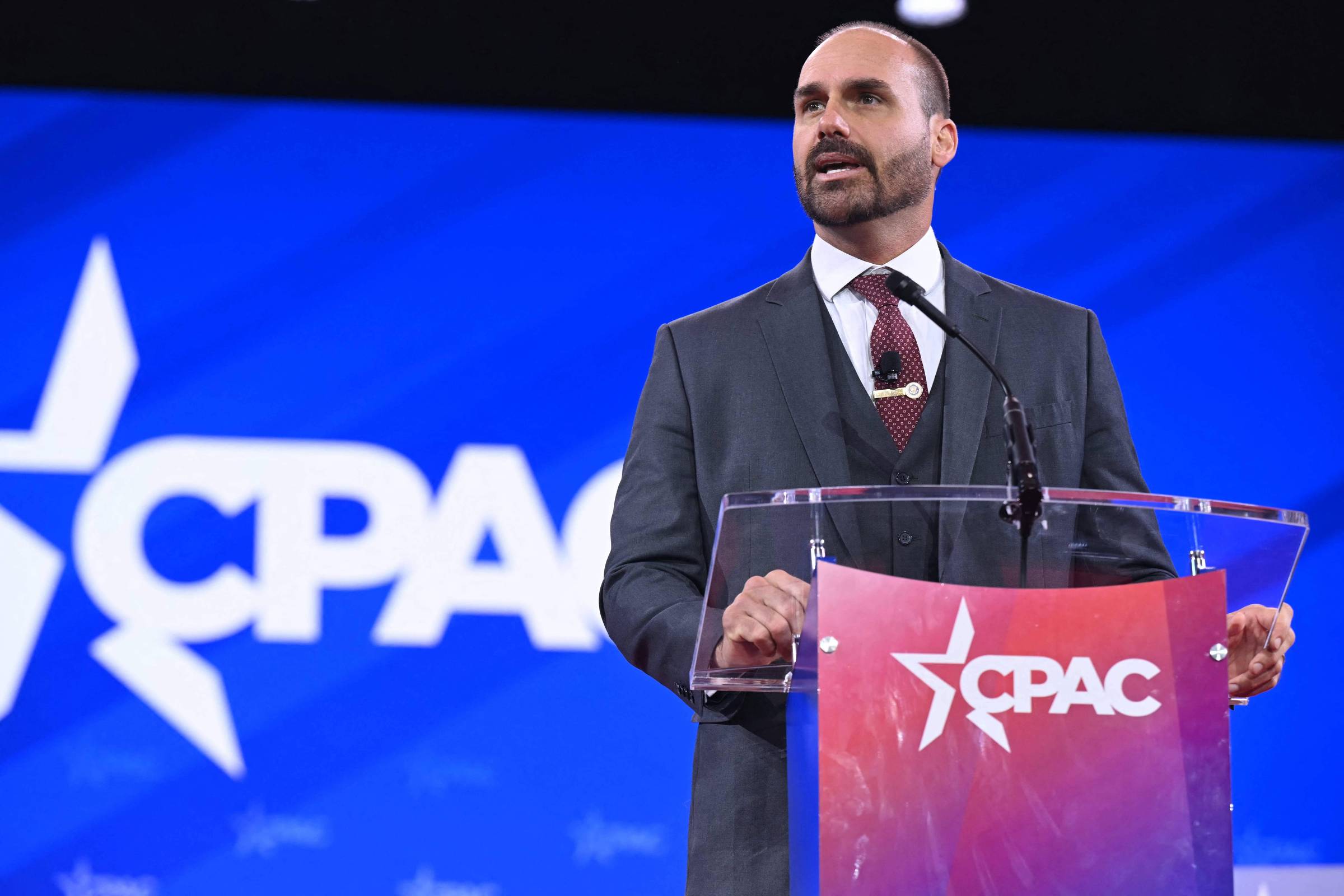 Um homem em um terno escuro está falando em um púlpito transparente com o logotipo do CPAC em destaque. O fundo é azul com o logotipo do CPAC em branco. O homem tem cabelo curto e barba bem aparada, e parece estar se dirigindo a uma audiência.