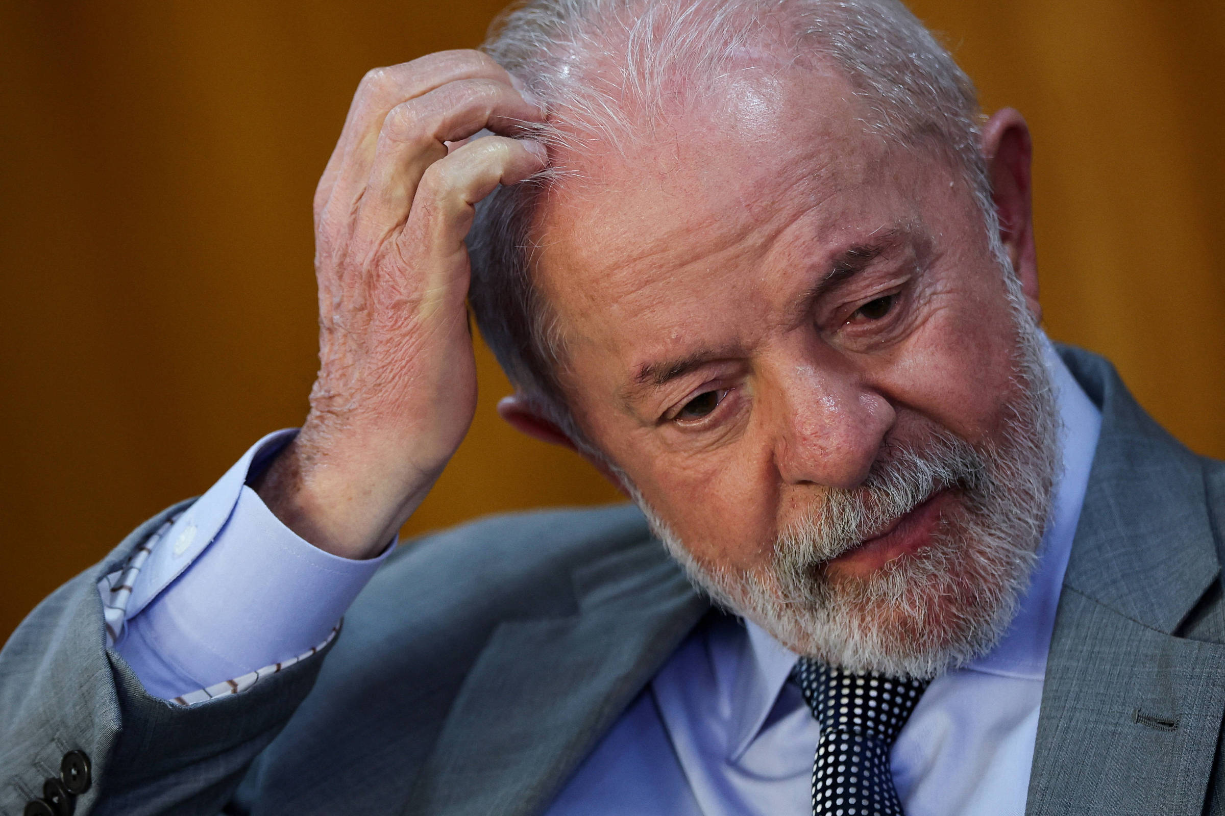 Um homem com cabelo grisalho e barba, vestindo um terno cinza e uma camisa azul clara, está com a mão na cabeça, parecendo pensativo ou preocupado. O fundo é de uma cor amarela suave, e a expressão facial do homem sugere reflexão ou incerteza.