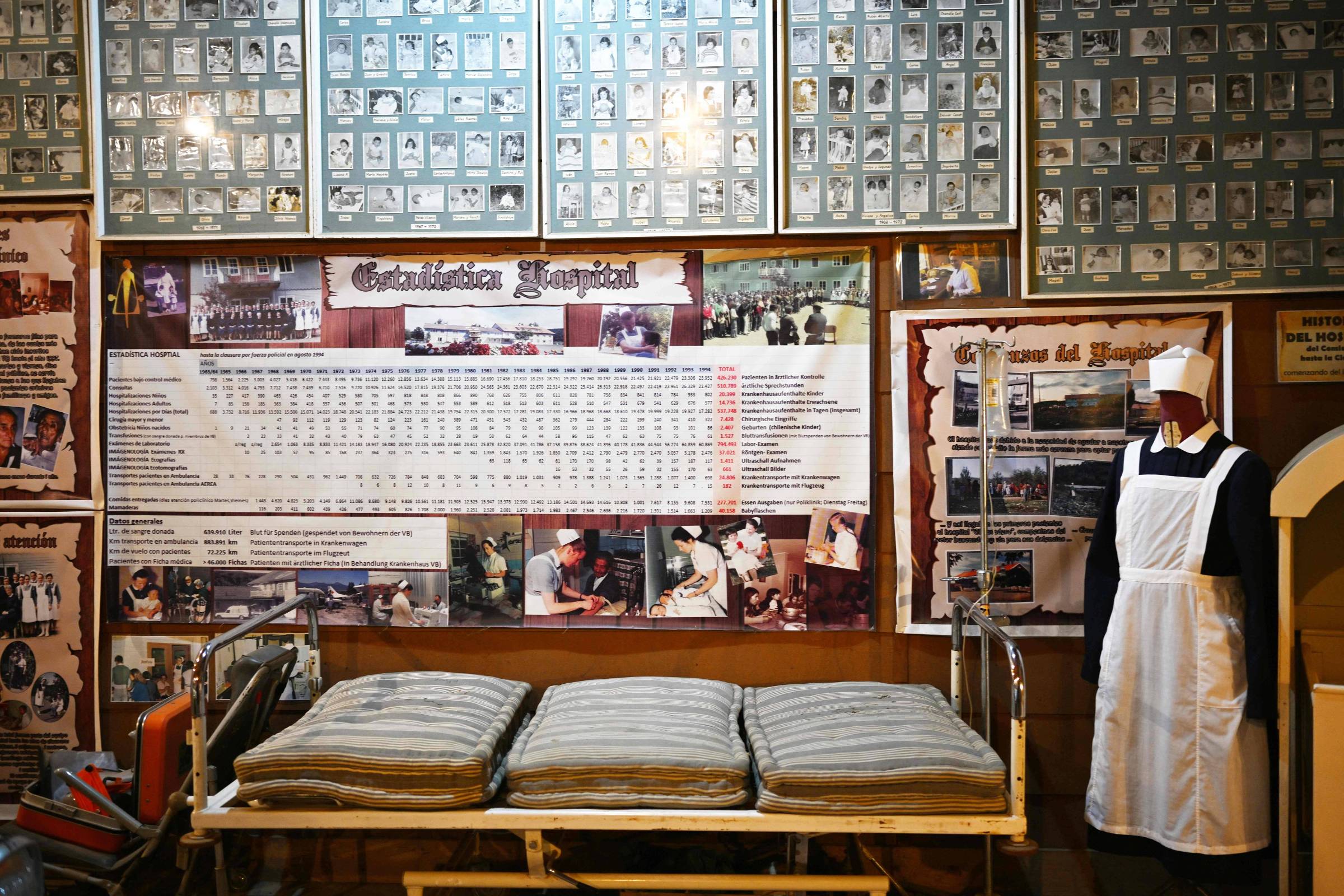 A imagem mostra uma exposição em um museu, com uma cama hospitalar antiga coberta por colchas listradas. Ao fundo, há uma parede repleta de quadros com fotografias de pessoas, possivelmente funcionários ou pacientes, e um grande painel informativo com dados e imagens relacionadas à história do hospital. À direita, uma manequim vestido com um uniforme de enfermeira, incluindo um avental e um chapéu.
