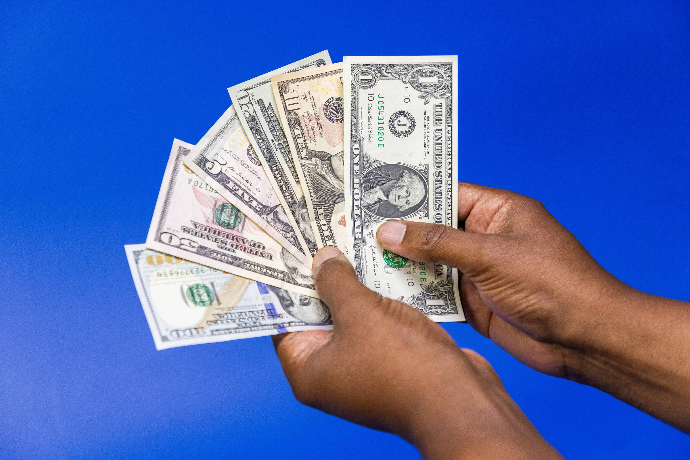 A imagem mostra uma mão segurando várias notas de dinheiro dos Estados Unidos, incluindo um dólar, cinco dólares, dez dólares, vinte dólares e cinquenta dólares, contra um fundo azul.