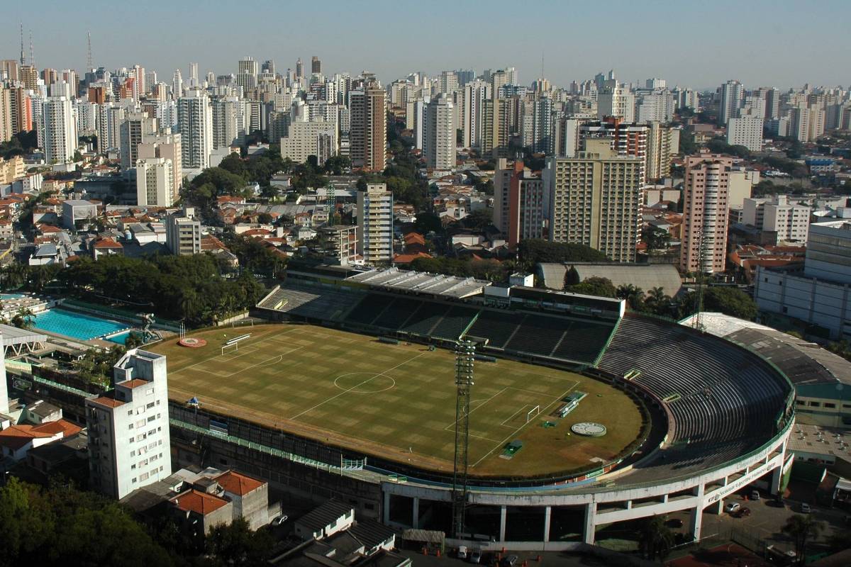A imagem mostra uma vista aérea de um estádio de futebol localizado em uma área urbana. O estádio possui um campo de grama e arquibancadas ao redor. Ao fundo, há uma densa paisagem urbana com muitos prédios altos e uma área residencial com casas de telhado vermelho. Também é visível uma piscina ao lado do estádio. O céu está claro e sem nuvens.