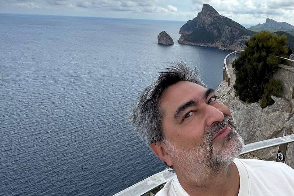 Zeca Camargo está tirando uma selfie em um local à beira-mar. Ao fundo, há uma vista do mar calmo e rochas à distância, com nuvens escuras no céu. O homem tem cabelo grisalho e usa uma camiseta branca com um design colorido. A paisagem é montanhosa e a atmosfera parece ser de um dia nublado.