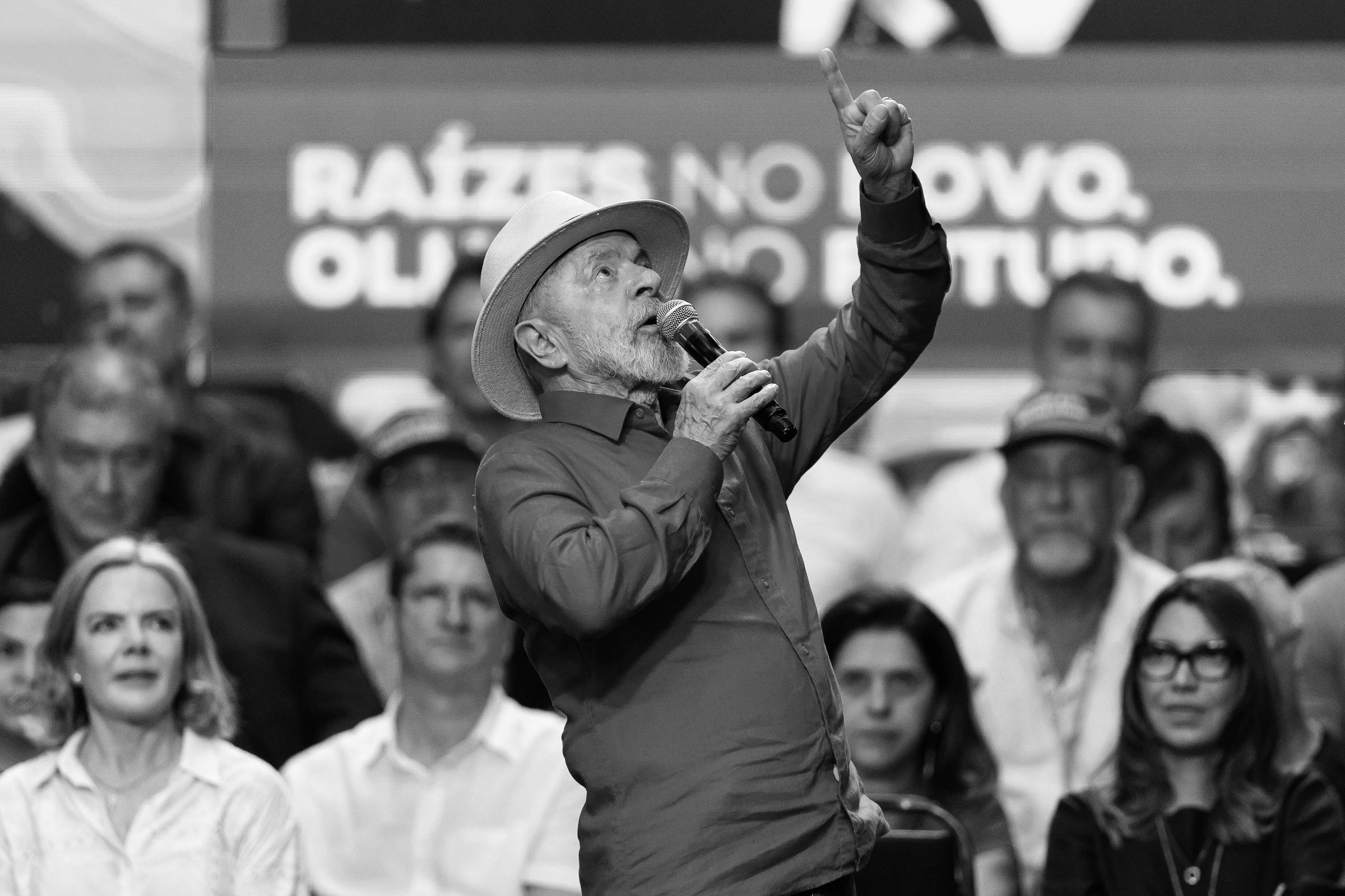 A imagem mostra um homem de cabelo grisalho, vestindo uma camisa vermelha e um chapéu claro, falando ao microfone em um evento político. Ele está gesticulando com a mão levantada. Ao fundo, há uma plateia com várias pessoas, algumas usando camisetas vermelhas. Um banner ao fundo contém o texto