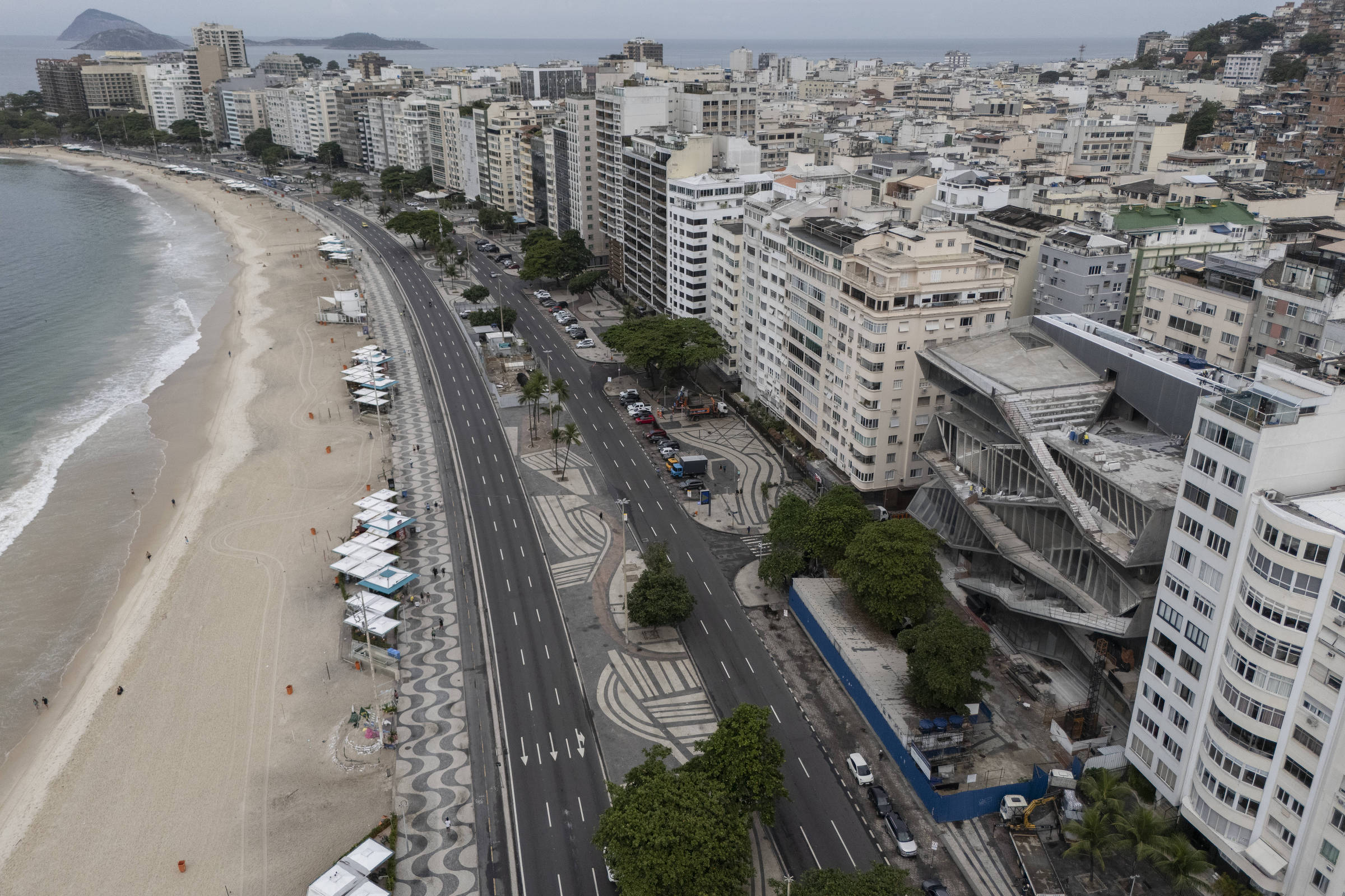 A imagem mostra uma vista aérea de uma praia com areia clara e um calçadão. À esquerda, há a praia e, à direita, uma avenida com prédios altos. O calçadão apresenta um padrão ondulado em preto e branco. Algumas barracas estão posicionadas ao longo da praia. O céu está nublado e há uma montanha ao fundo, além de várias pessoas visíveis na praia e na avenida.