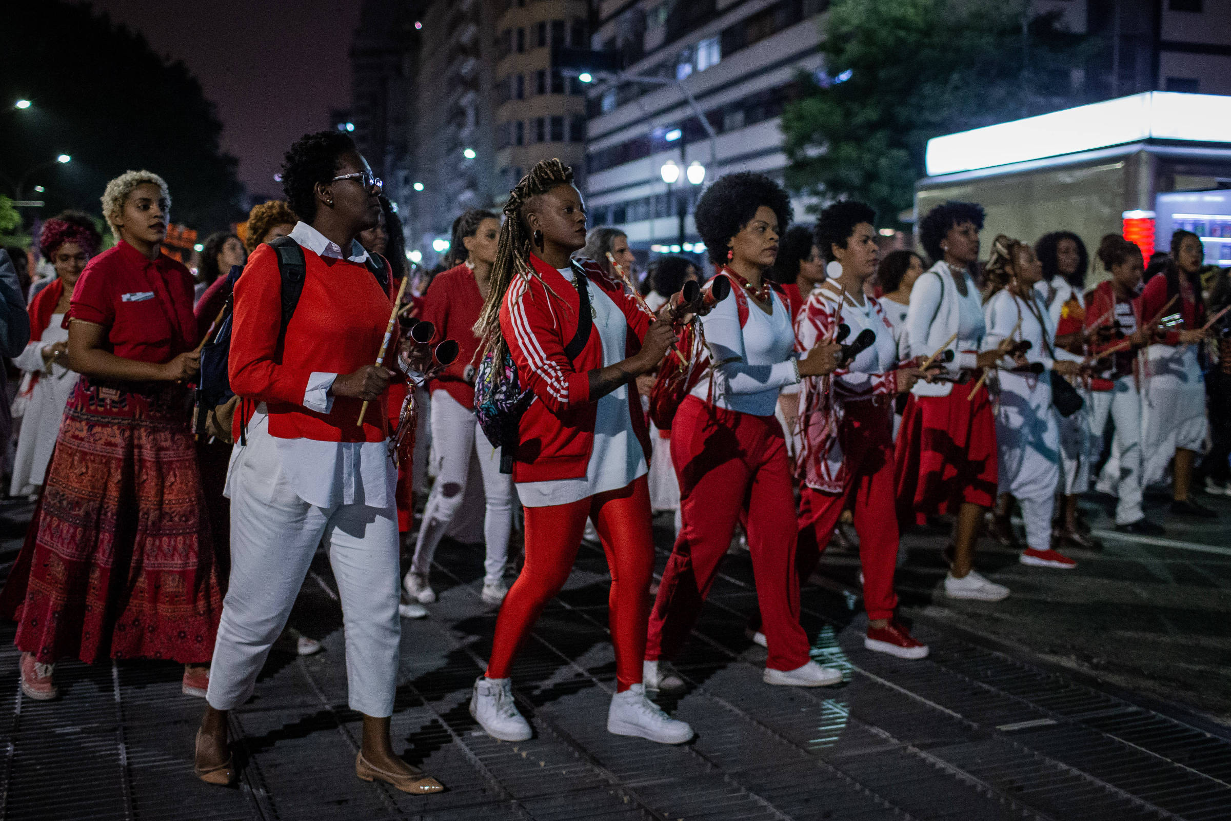 A imagem mostra um grupo de mulheres marchando em uma rua à noite. Elas estão vestidas em roupas brancas e vermelhas, algumas segurando instrumentos musicais. O ambiente é urbano, com prédios ao fundo e iluminação de rua visível. A atmosfera parece ser de celebração ou protesto, com as participantes demonstrando união e energia.