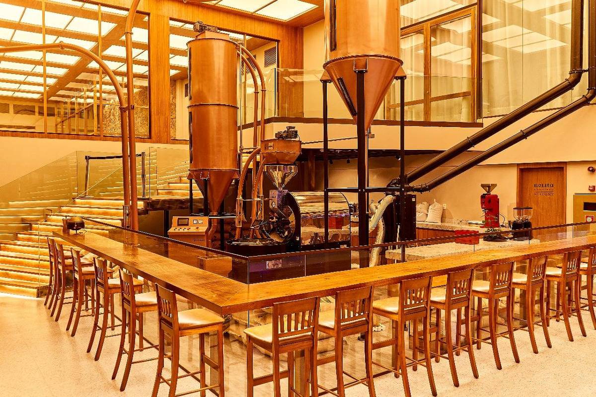 A imagem mostra o interior de uma cafeteria moderna, com um grande balcão de madeira e várias cadeiras ao redor. No fundo, há um sistema de tubulações de cobre e equipamentos de café, incluindo um moinho e um torrador. As paredes são de vidro, permitindo a entrada de luz natural, e o ambiente é bem iluminado e acolhedor.