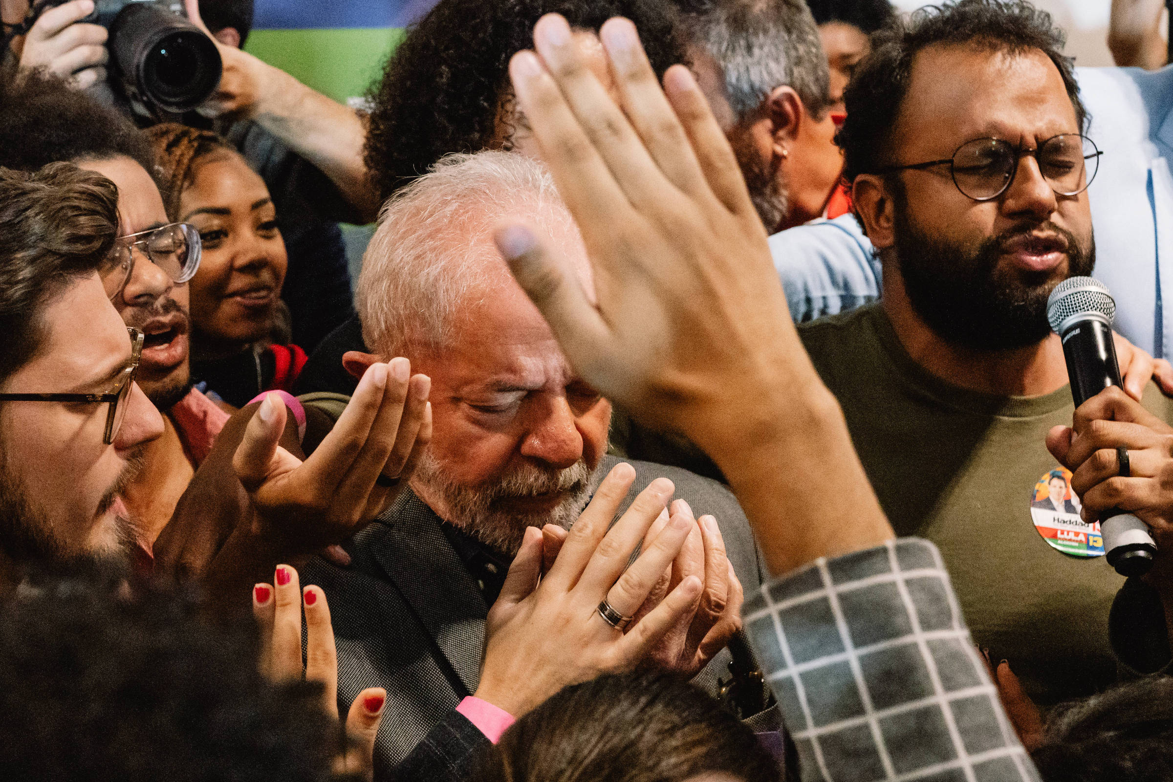 A imagem mostra um grupo de pessoas em um evento político, com destaque para um homem de cabelos grisalhos e barba, que parece emocionado, com os olhos fechados e as mãos unidas em um gesto de oração ou agradecimento. Ao seu redor, várias mãos estão levantadas, algumas tocando-o, enquanto outras seguram microfones e câmeras. O ambiente é de intensa interação e apoio emocional.