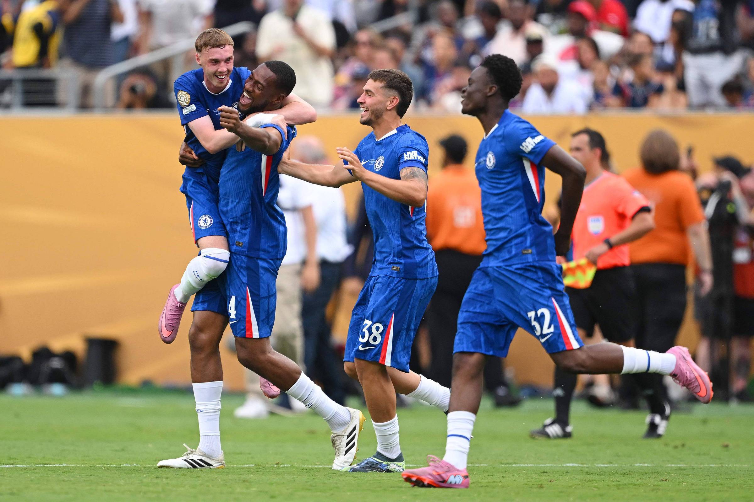 Quatro jogadores de futebol estão celebrando em campo. Um jogador está sendo carregado nas costas de outro, enquanto os outros dois jogadores estão ao redor, sorrindo e comemorando. Todos estão vestindo uniformes azuis com detalhes brancos. O fundo mostra uma multidão de torcedores e outros jogadores, criando um ambiente animado de jogo.