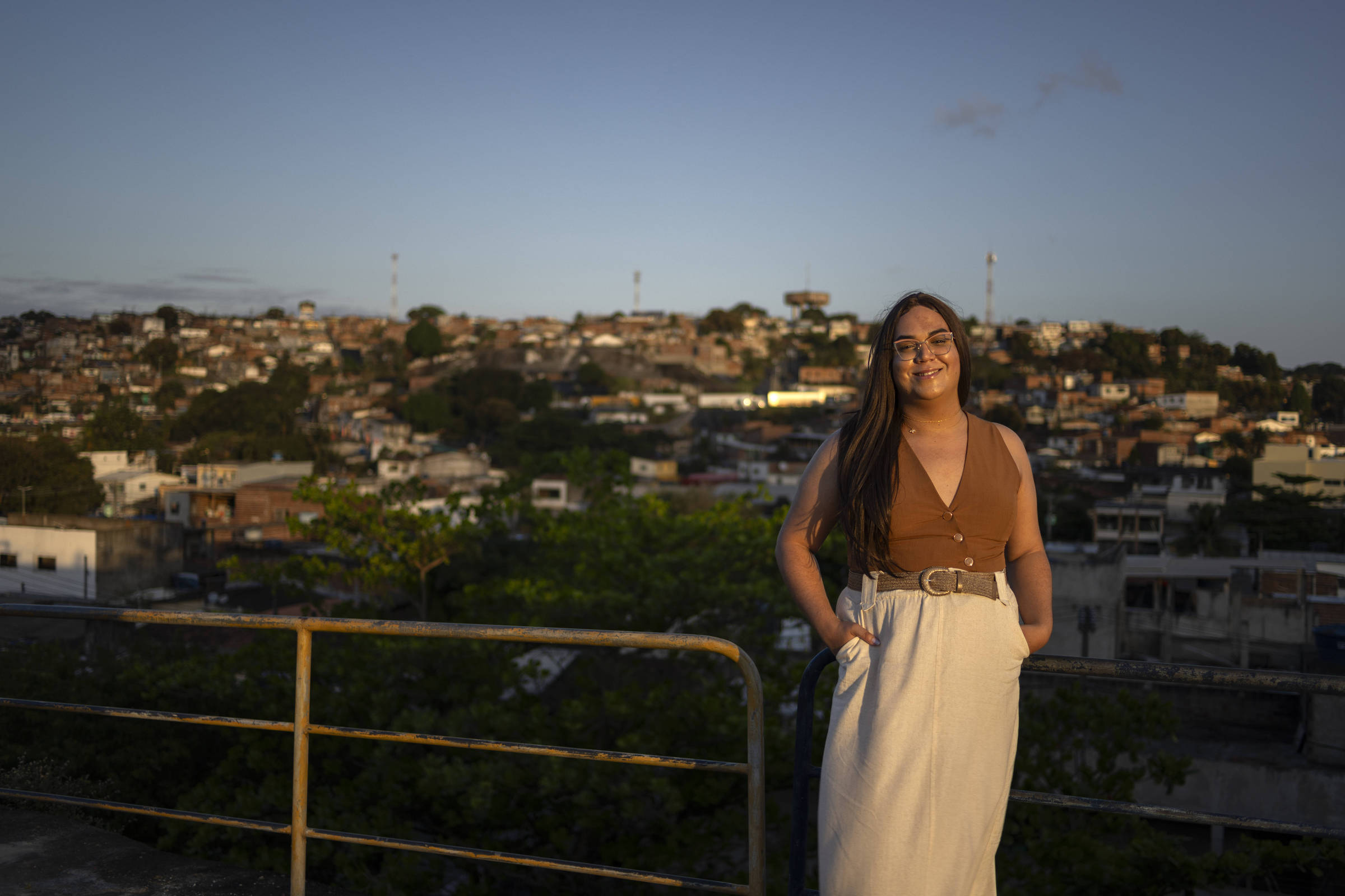Uma mulher de cabelos compridos, blusa marrom e saia branca posa em frente a uma cidade