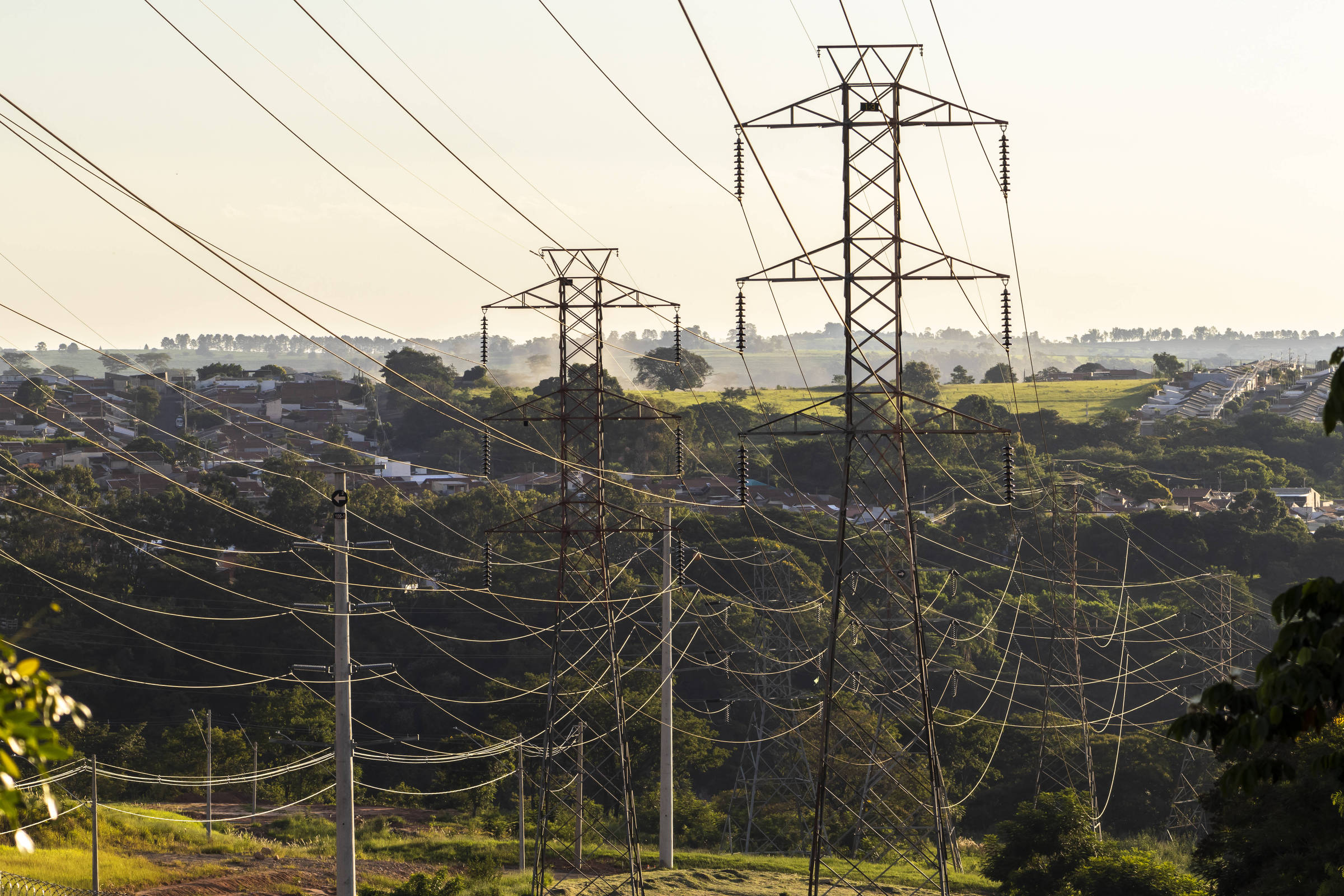 A imagem mostra várias torres de transmissão de energia elétrica em uma área rural. As torres estão alinhadas em uma linha, com fios de alta tensão se estendendo entre elas. Ao fundo, há uma paisagem verde com árvores e uma pequena área urbana visível. O céu está claro, indicando um dia ensolarado.