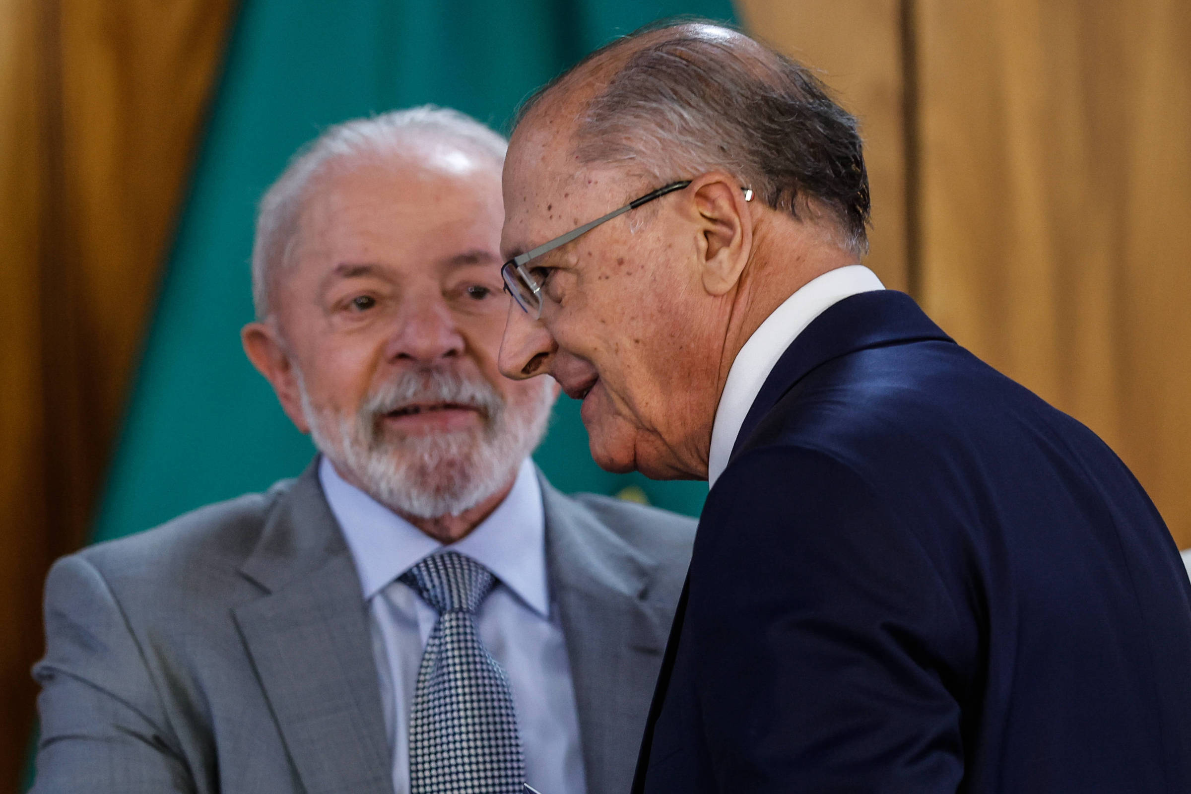 A imagem mostra dois homens em um evento formal. O homem à esquerda, com cabelo grisalho e barba, está sorrindo e olhando para o homem à direita, que está de perfil e usa um terno escuro. O fundo é composto por uma cortina verde e uma parede de madeira.
