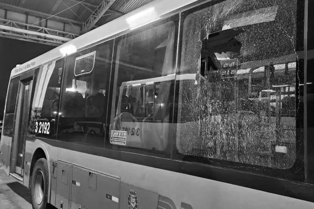 A imagem mostra um ônibus estacionado em um local coberto, com um vidro lateral quebrado. O vidro está estilhaçado, e a parte interna do ônibus é visível, com assentos e outros elementos do interior. O ônibus é de cor amarela e cinza, e há um número de identificação visível na lateral, que é 2102.