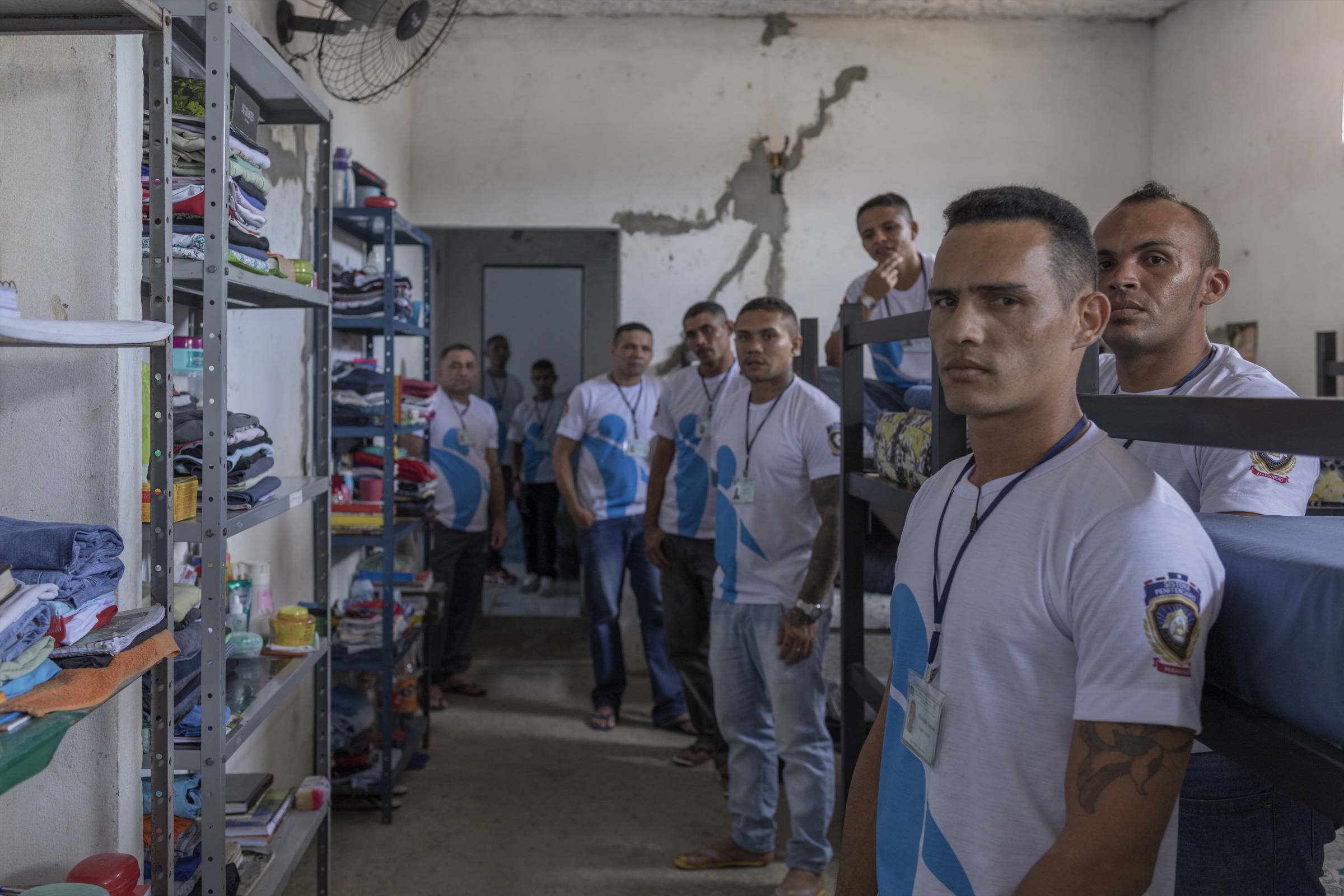 A imagem mostra um grupo de homens em um ambiente interno, possivelmente uma cela ou dormitório. Eles estão alinhados em uma fileira, todos usando camisetas brancas com um desenho azul. O ambiente é simples, com prateleiras ao fundo contendo roupas e outros itens pessoais. As paredes estão desgastadas e há um ventilador de teto visível. Os homens parecem estar em uma atividade organizada, com expressões sérias.