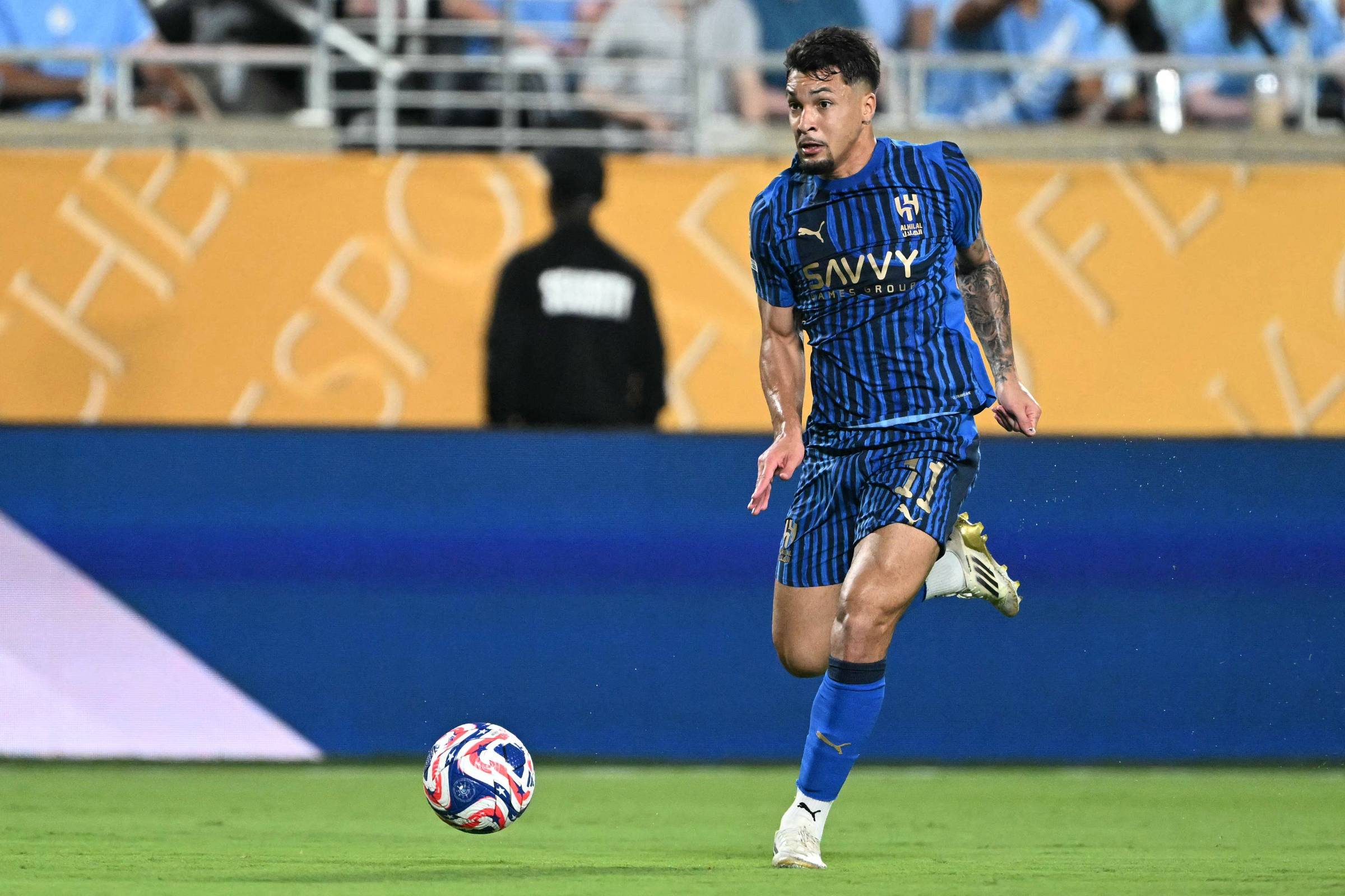 Um jogador de futebol está correndo em campo, vestindo um uniforme azul listrado com detalhes em preto. Ele está em movimento, com a bola de futebol próxima aos pés. Ao fundo, há uma arquibancada com torcedores e uma faixa amarela com texto visível. Um segurança pode ser visto em pé, ao fundo, com um uniforme escuro.