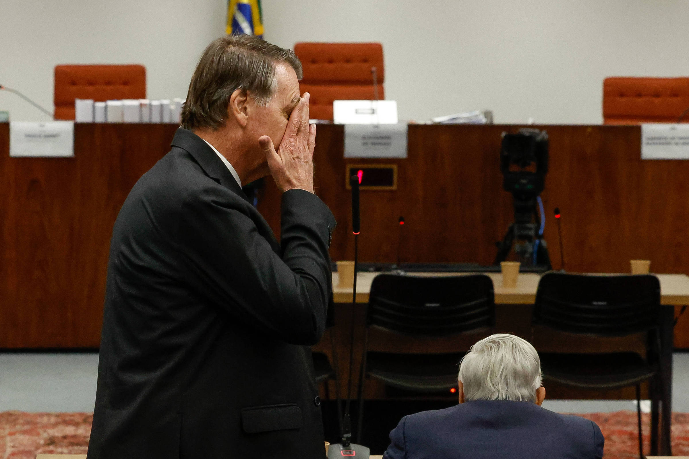 A imagem mostra um homem de terno escuro, com cabelo curto e grisalho, em pé, com a mão no rosto, aparentando estar pensativo ou preocupado. Ao fundo, há uma mesa de tribunal com cadeiras e uma bandeira do Brasil visível. O ambiente parece ser uma sala de audiência, com um cenário formal e organizado.