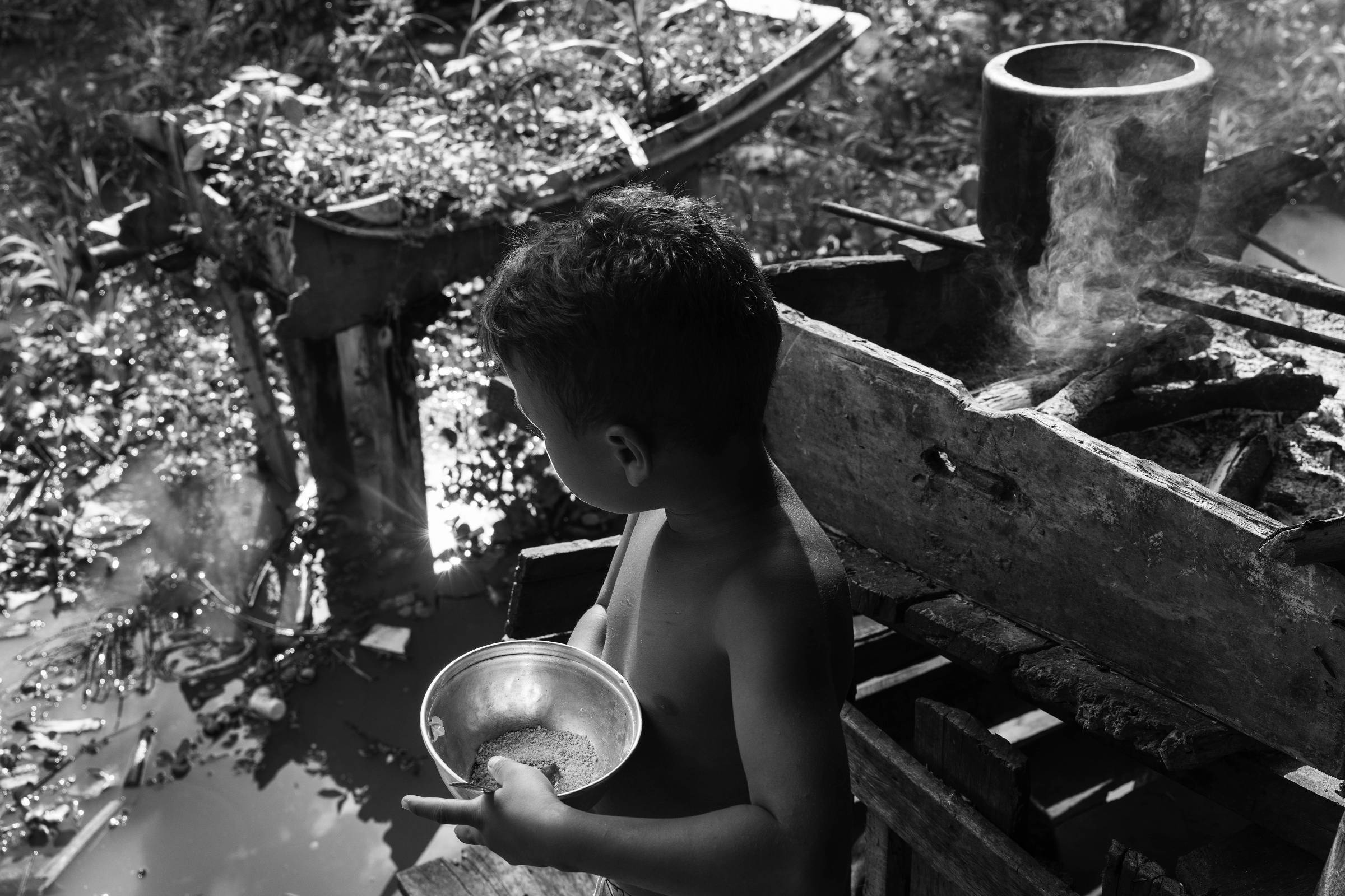 A imagem mostra uma criança sem camisa, de costas, segurando uma tigela de metal. Ao fundo, há uma fogueira com fumaça saindo e um recipiente de metal em cima dela. O ambiente é rural, com vegetação ao redor e um barco parcialmente visível. O chão está coberto por água e plantas.