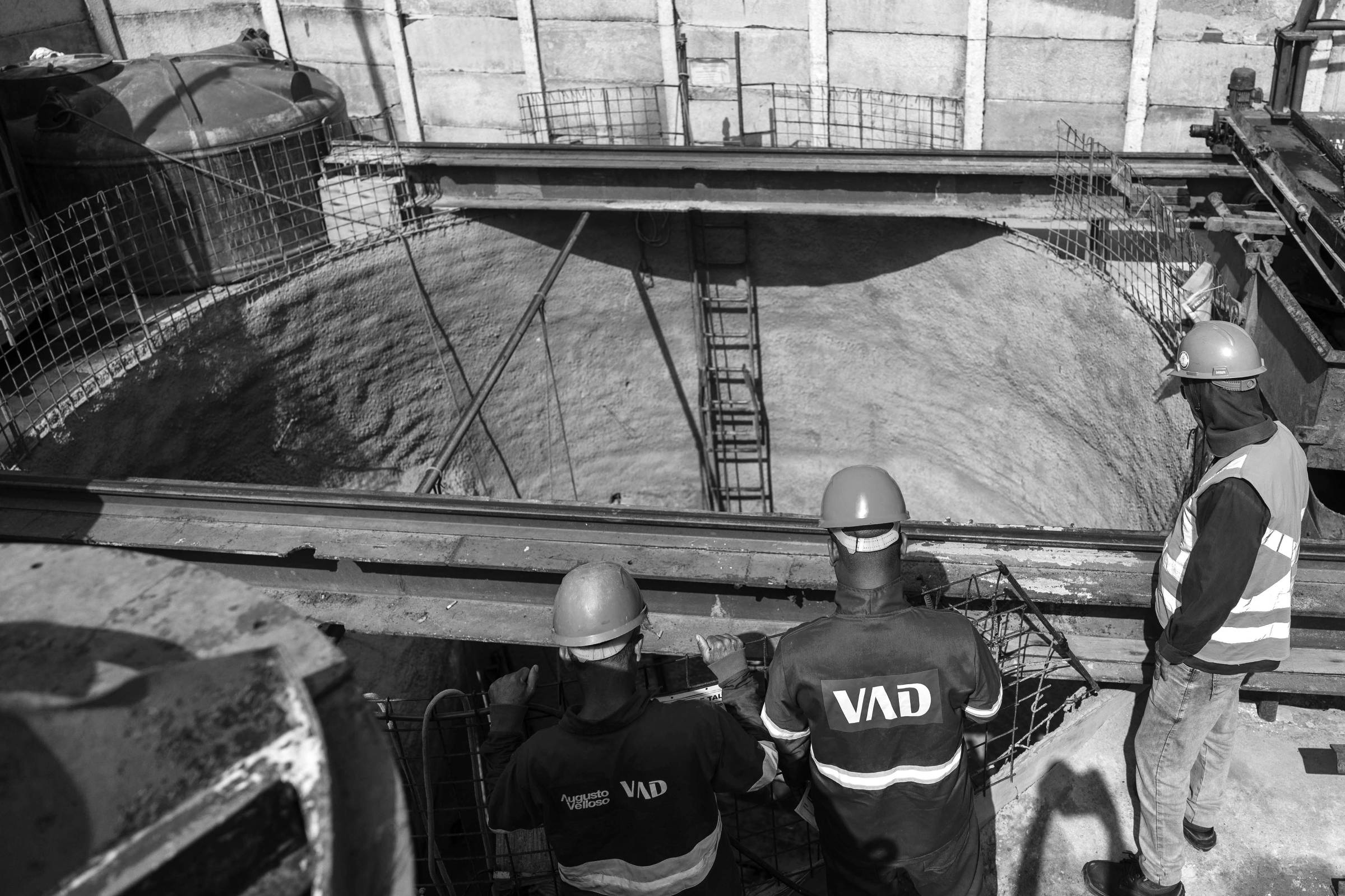 A imagem mostra um grupo de trabalhadores em um canteiro de obras, observando uma grande escavação circular. Há uma estrutura de metal acima da escavação, com uma escada visível. Os trabalhadores usam capacetes de segurança em cores diferentes: um amarelo, um verde e um azul. Um deles está usando um colete laranja refletivo. Ao fundo, há uma parede de contenção e alguns materiais de construção.