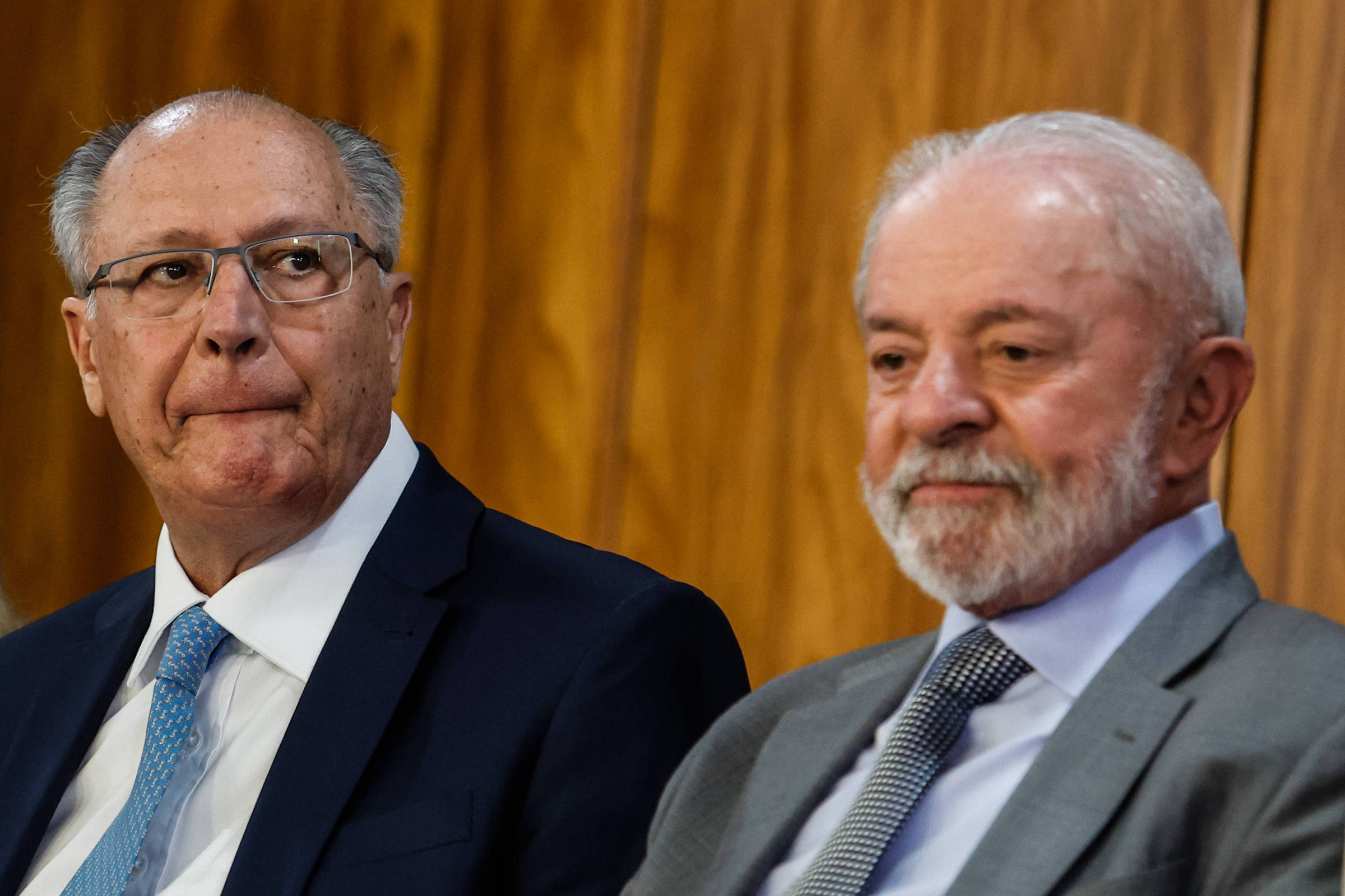 A imagem mostra dois homens sentados lado a lado em um ambiente formal. O homem à esquerda, com cabelo grisalho e óculos, está olhando para o lado com uma expressão pensativa. O homem à direita, com barba e cabelo grisalho, está sorrindo levemente. O fundo é de madeira, sugerindo um ambiente de reunião ou conferência.