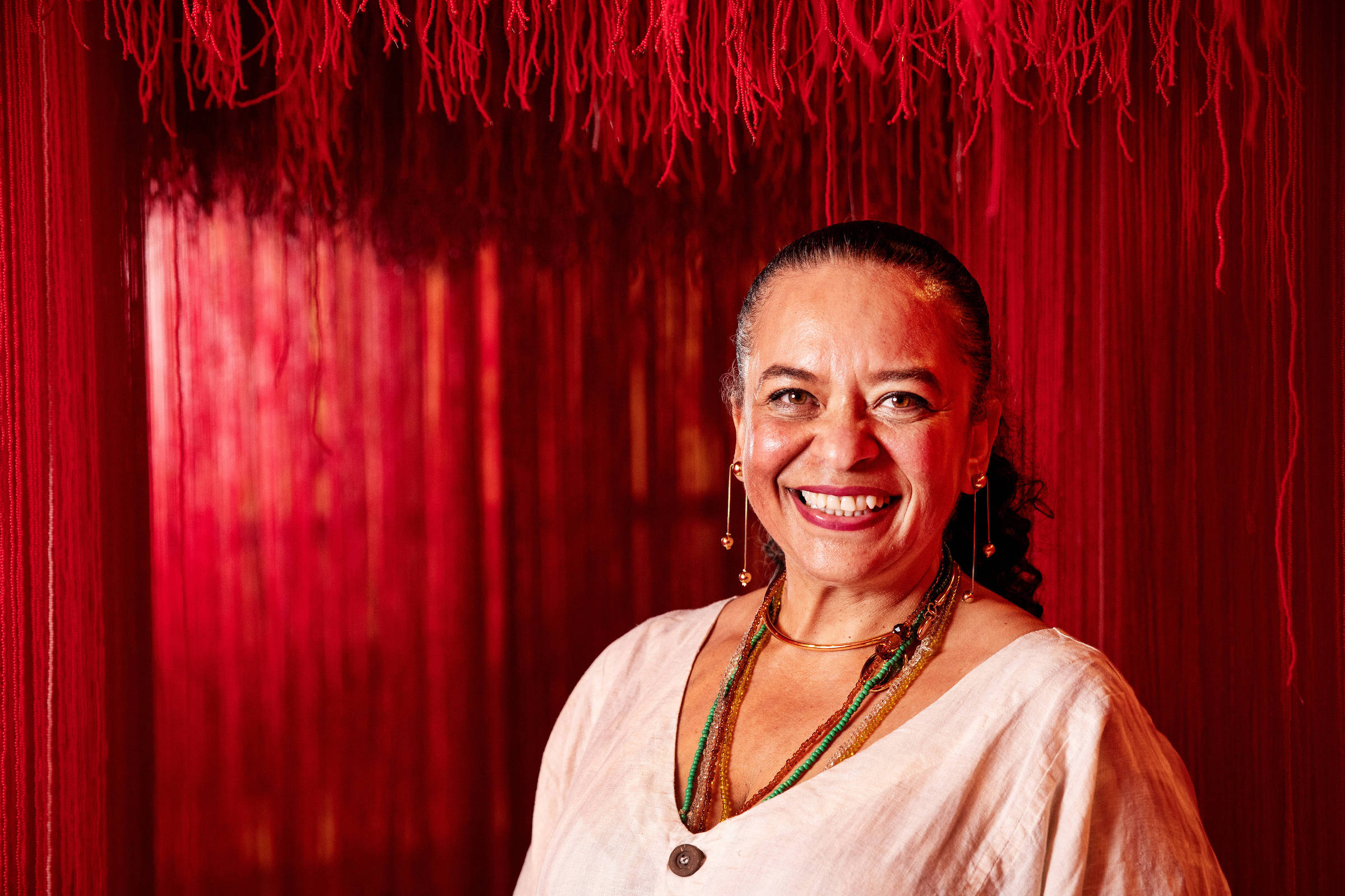 A imagem mostra uma mulher sorridente em um ambiente com fundo vermelho. Ela usa uma blusa clara e colares coloridos. O cenário é decorado com fios vermelhos que criam um efeito visual interessante.
