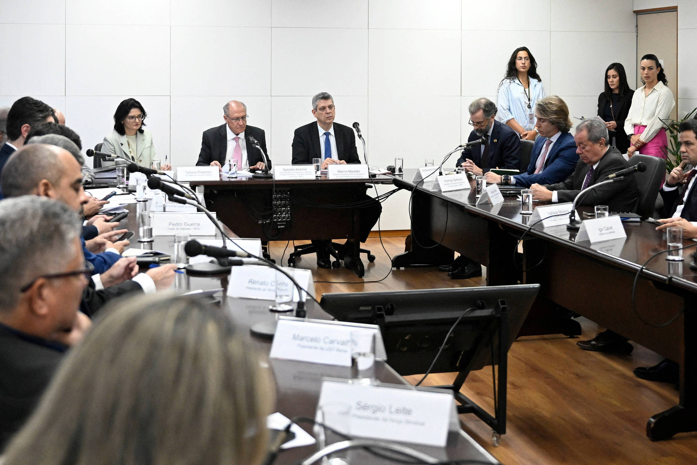 A imagem mostra uma reunião em uma sala com uma mesa grande em formato oval. Várias pessoas estão sentadas ao redor da mesa, algumas com laptops e documentos. No centro, há um grupo de homens e mulheres em trajes formais, discutindo. Ao fundo, algumas pessoas estão em pé, observando. A sala tem paredes brancas e um piso de madeira.