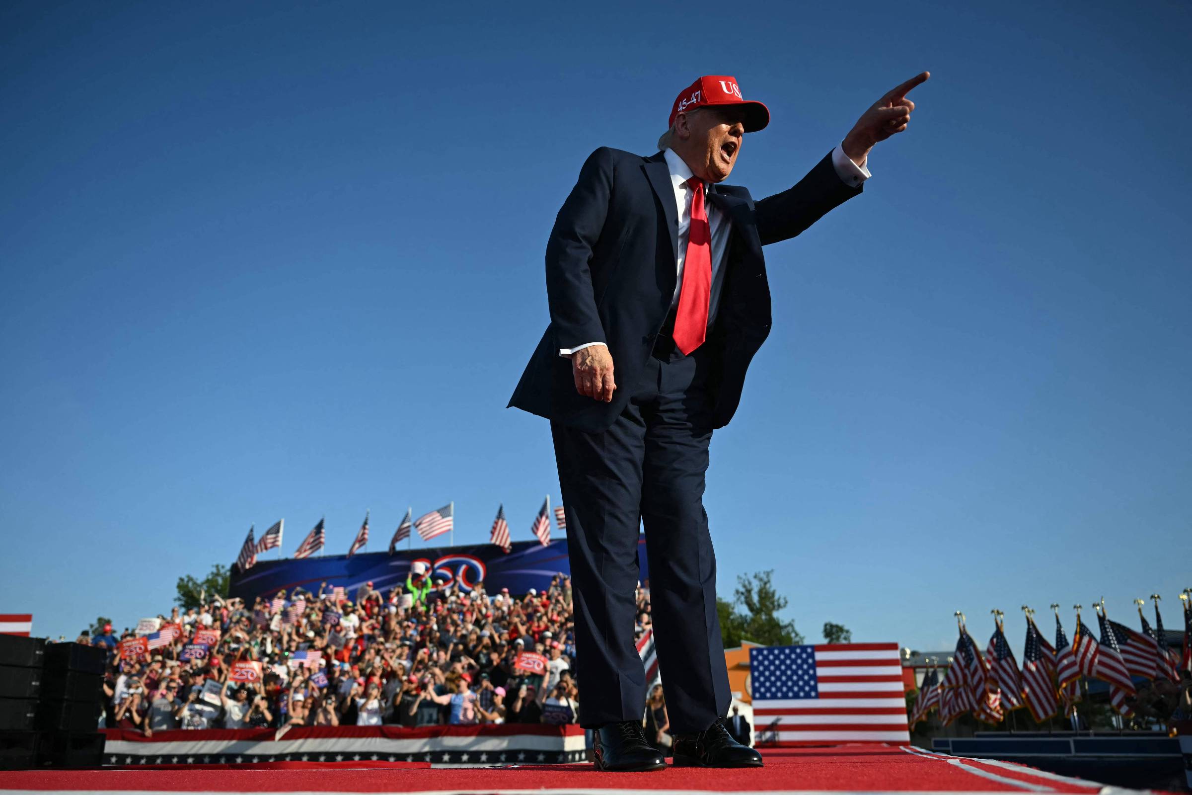 Um homem em um terno escuro e gravata vermelha está em um palco, apontando para a frente. Ele usa um boné vermelho e está cercado por uma multidão de pessoas que seguram bandeiras. Ao fundo, há uma grande quantidade de bandeiras americanas e um céu azul claro.