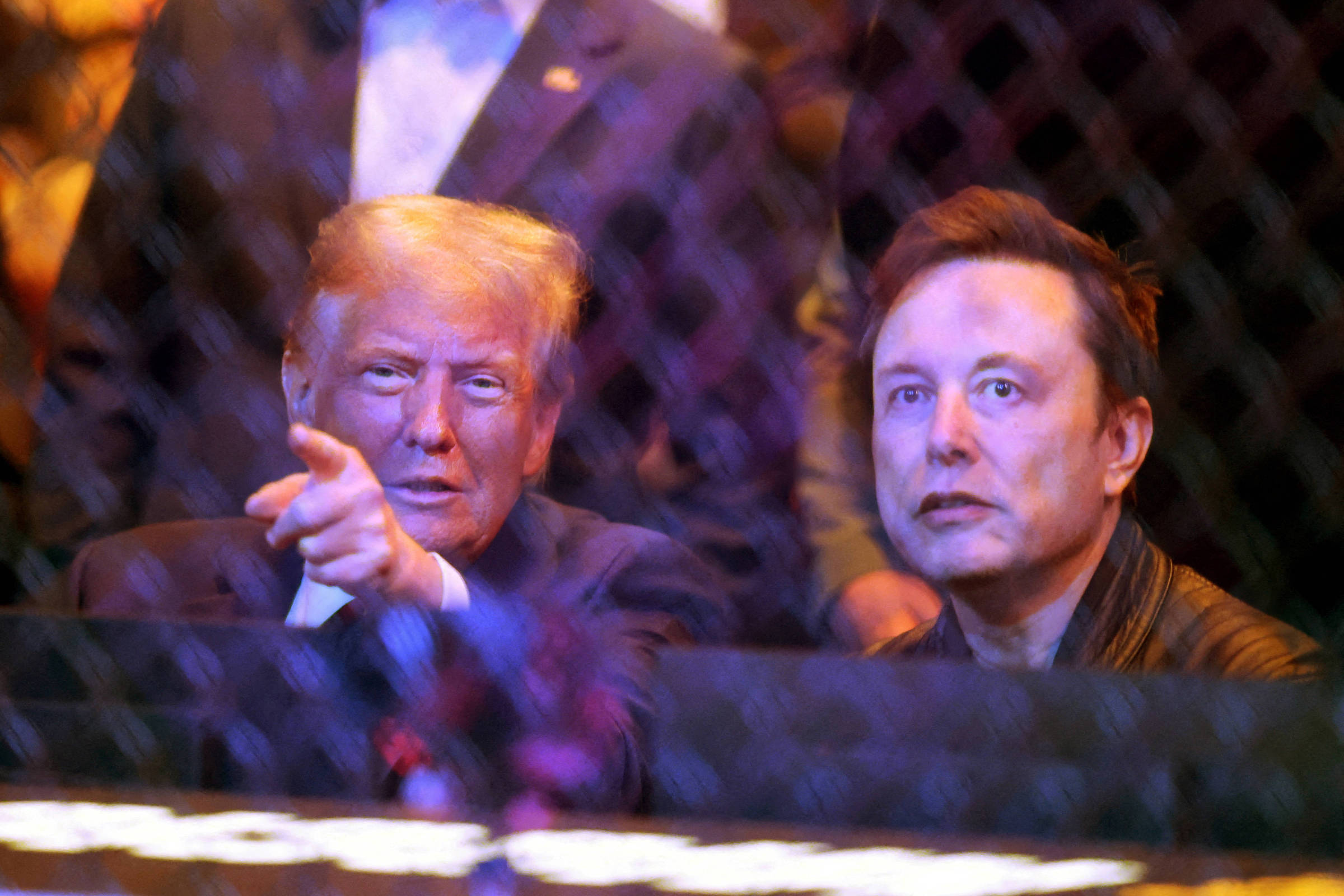 A imagem mostra duas pessoas sentadas em um evento, observando algo à frente. Uma delas está apontando com o dedo, enquanto a outra parece atenta. O ambiente é iluminado e há uma grade de proteção visível em primeiro plano.
