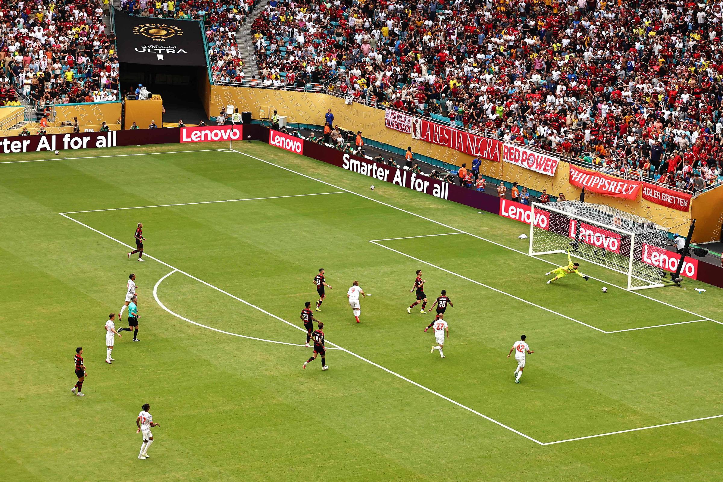 A imagem mostra um jogo de futebol em um estádio lotado. No campo, jogadores de duas equipes estão em ação, com um jogador prestes a chutar a bola em direção ao gol. O goleiro está se preparando para defender. Ao fundo, a torcida está animada, com muitas pessoas nas arquibancadas. Várias placas publicitárias estão visíveis ao redor do campo, incluindo uma da Lenovo.