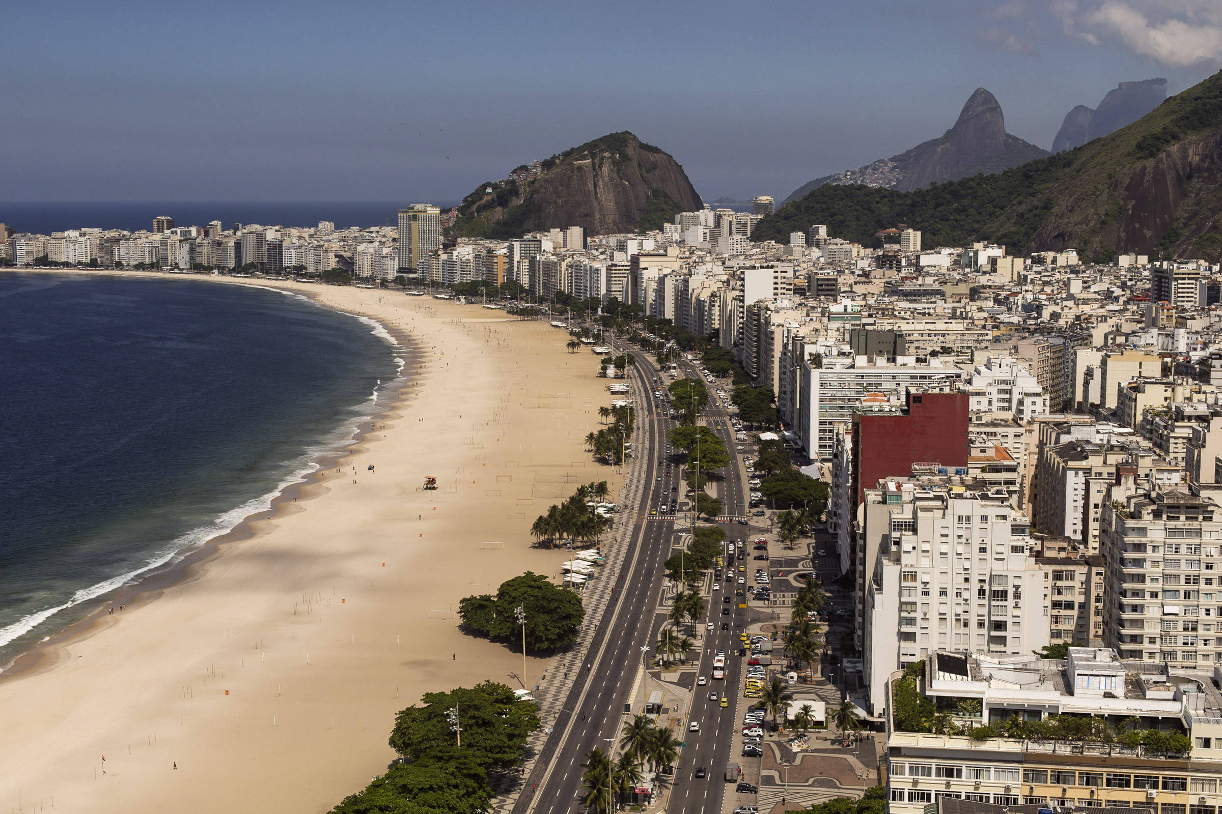 A imagem mostra uma vista panorâmica da Praia de Copacabana, no Rio de Janeiro. A praia é extensa, com areia clara e ondas do mar. Ao fundo, há uma linha de prédios altos e modernos, e montanhas características da região. A estrada que acompanha a praia é visível, com algumas árvores e veículos. O céu está claro, com algumas nuvens.