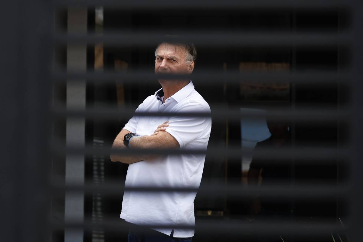 Um homem está em pé, com os braços cruzados, olhando para frente. Ele usa uma camisa branca e está posicionado atrás de uma grade ou persiana, que cria um efeito de linhas verticais na imagem. O fundo é desfocado, mas parece haver uma área externa iluminada.