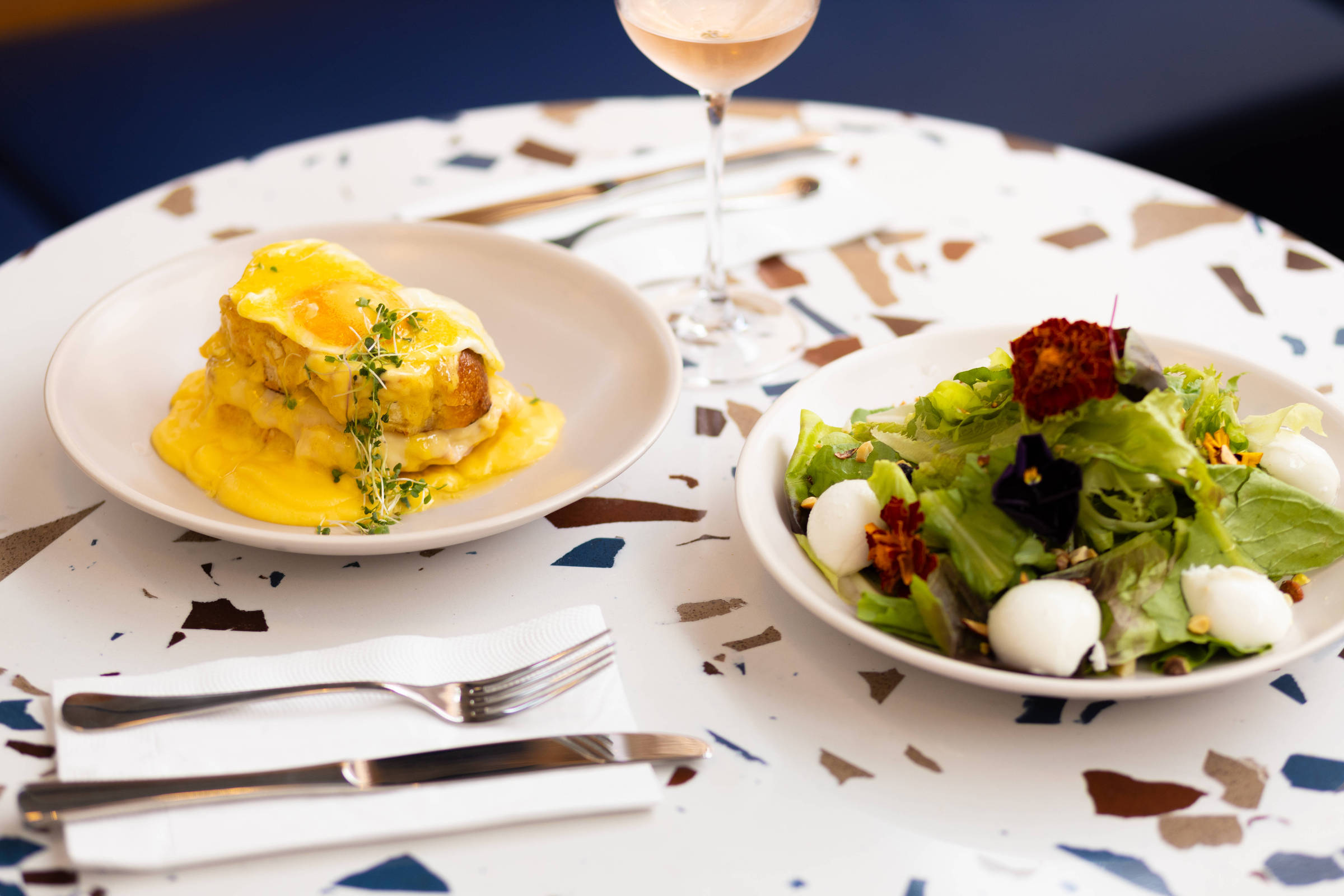 A imagem mostra uma mesa com um prato principal e uma salada. O prato principal, à esquerda, contém uma toast coberta com um molho amarelo e decorada com um raminho de tomilho. À direita, há uma salada composta por folhas verdes, flores comestíveis e chips de vegetais. Ao fundo, há um copo de vinho rosé e a mesa é decorada com um padrão de mosaico em várias cores.