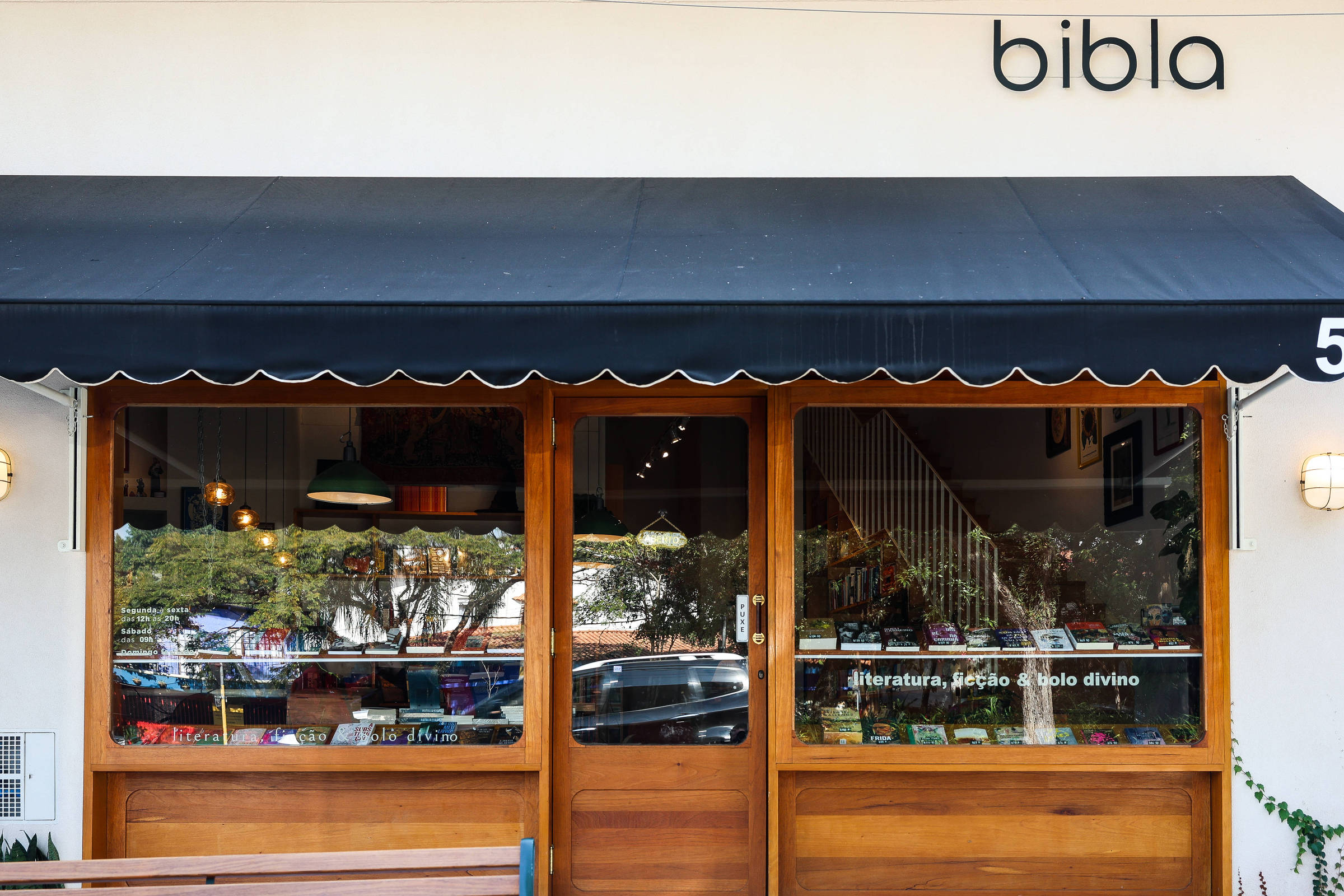 Fachada da livraria Bibla com porta e janelas de madeira, toldo preto e número 58 à direita. Na vitrine, livros expostos e texto