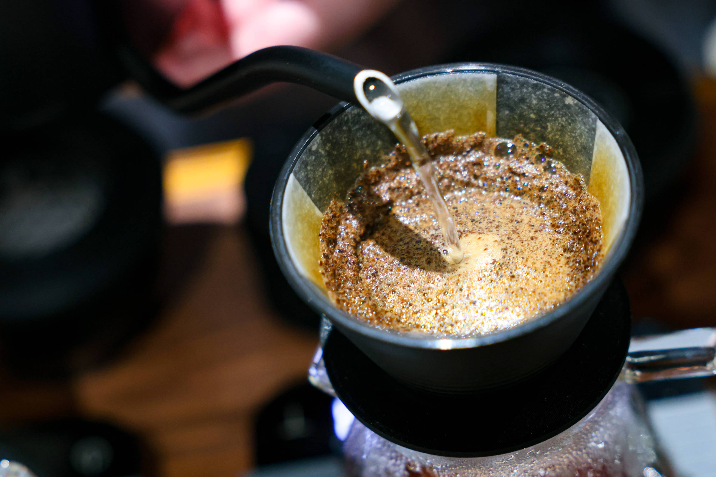 Água quente é despejada lentamente sobre pó de café em filtro de papel dentro de um coador preto. O café está em processo de extração, com espuma e borbulhas visíveis na superfície.