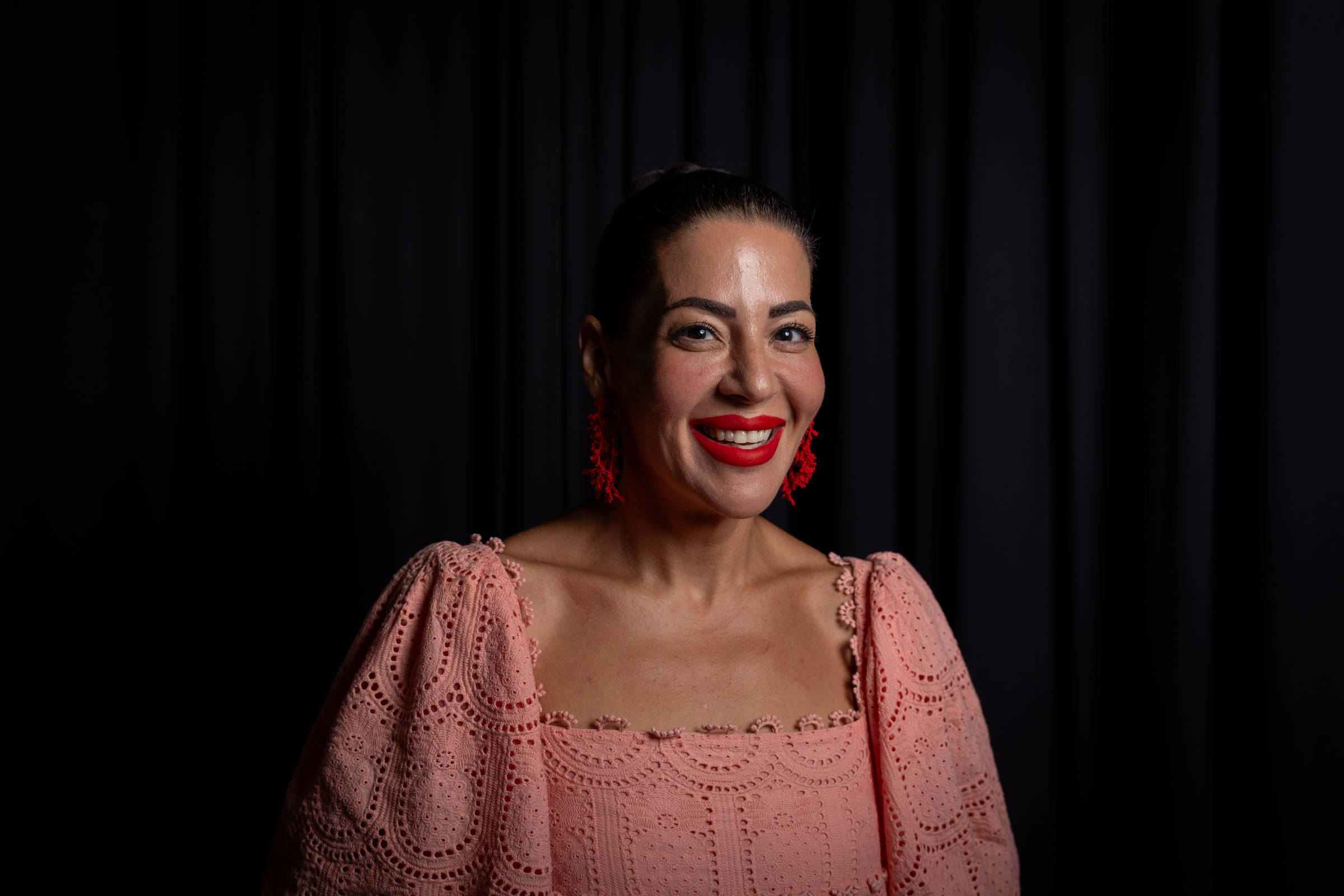 A imagem mostra uma mulher sorridente com cabelo preso em um coque. Ela usa uma blusa rosa com detalhes em crochê e brincos grandes e vermelhos. O fundo é preto, criando um contraste com sua roupa e expressão.