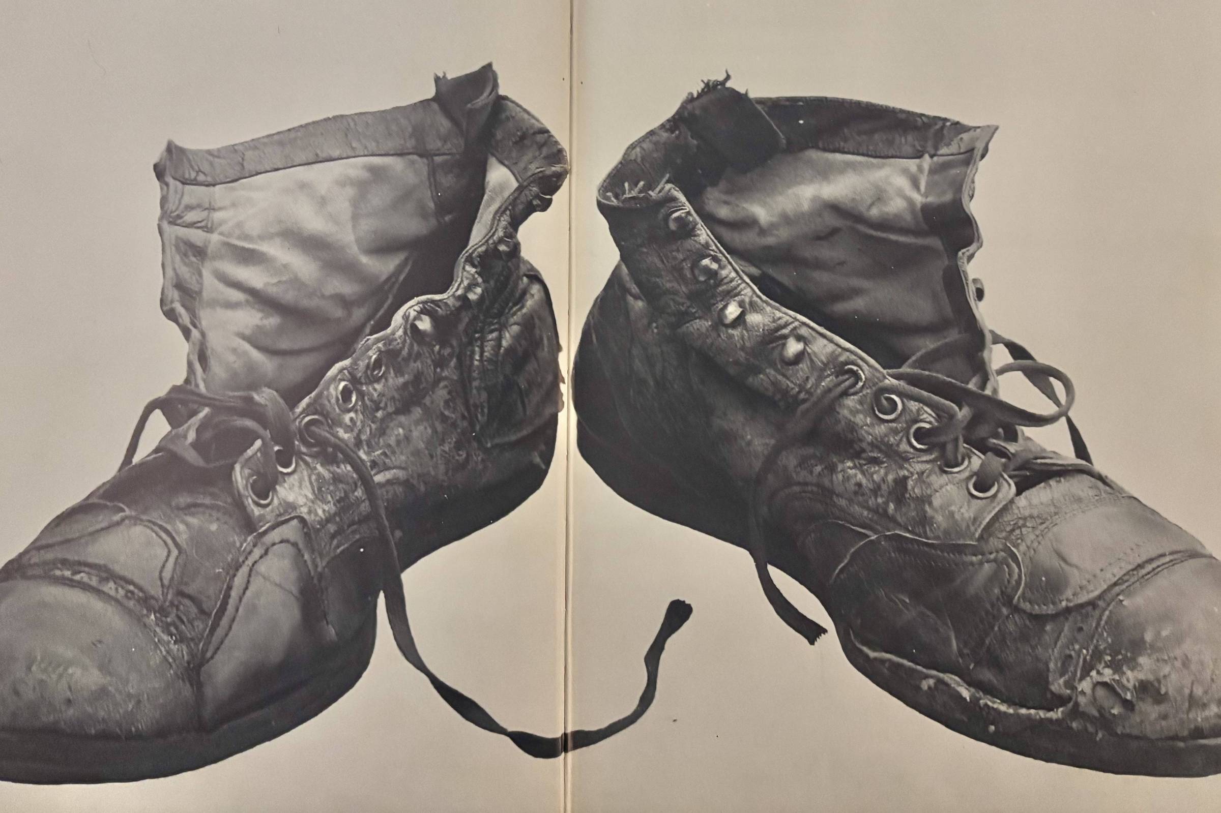 Par de botas de couro pretas muito gastas e amarradas, com sinais visíveis de uso intenso, incluindo vincos, rasgos e desgaste na sola e na parte superior.