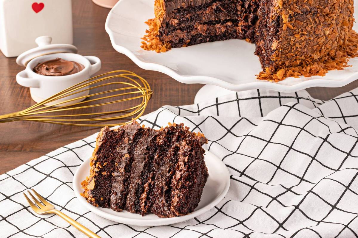 A imagem mostra um bolo de chocolate com várias camadas, coberto com flocos crocantes. O bolo está em um prato branco, com uma fatia servida ao lado em um prato menor. Ao fundo, há um recipiente rosa com a inscrição