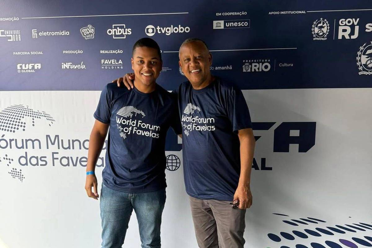 Dois homens posam lado a lado com os braços nos ombros um do outro, ambos vestindo camisetas azuis com a inscrição