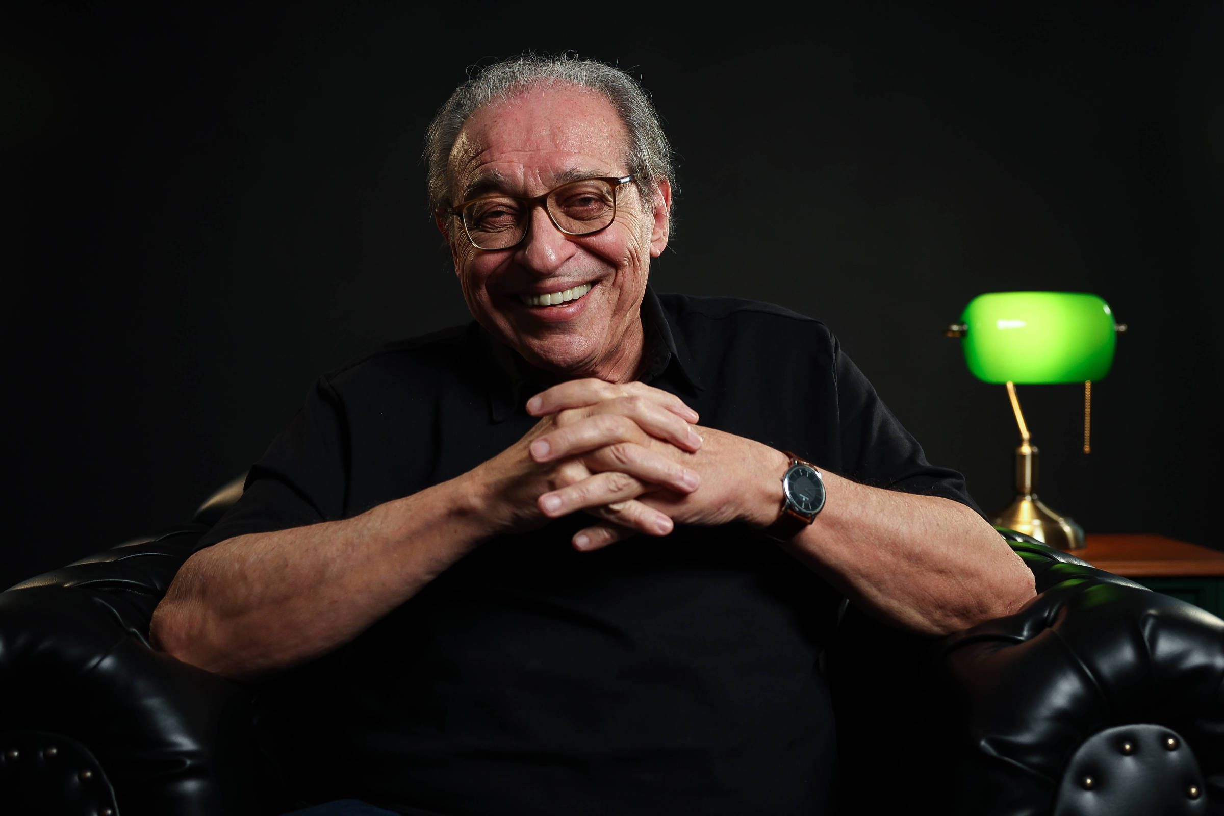 Um homem de cabelos grisalhos e óculos, sorrindo enquanto está sentado em um sofá de couro preto. Ele está usando uma camiseta preta e tem as mãos entrelaçadas. Ao fundo, há uma lâmpada verde acesa sobre uma mesa, criando um ambiente acolhedor e iluminado.