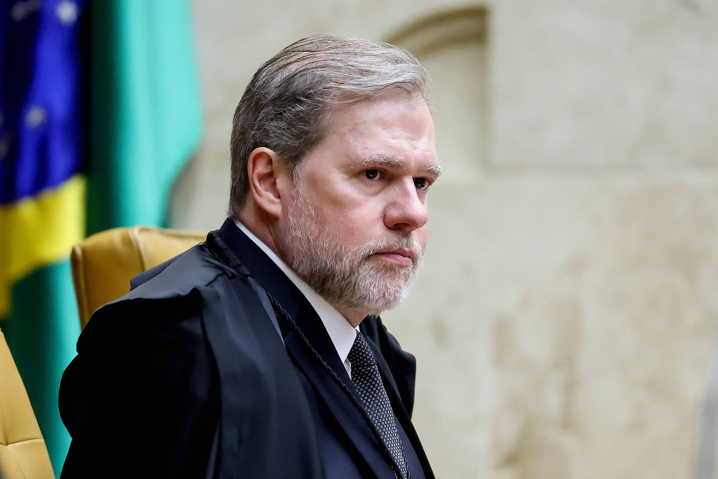 Ministro do Supremo Tribunal Federal sentado em cadeira oficial, vestindo toga preta e gravata escura, com bandeiras desfocadas ao fundo.