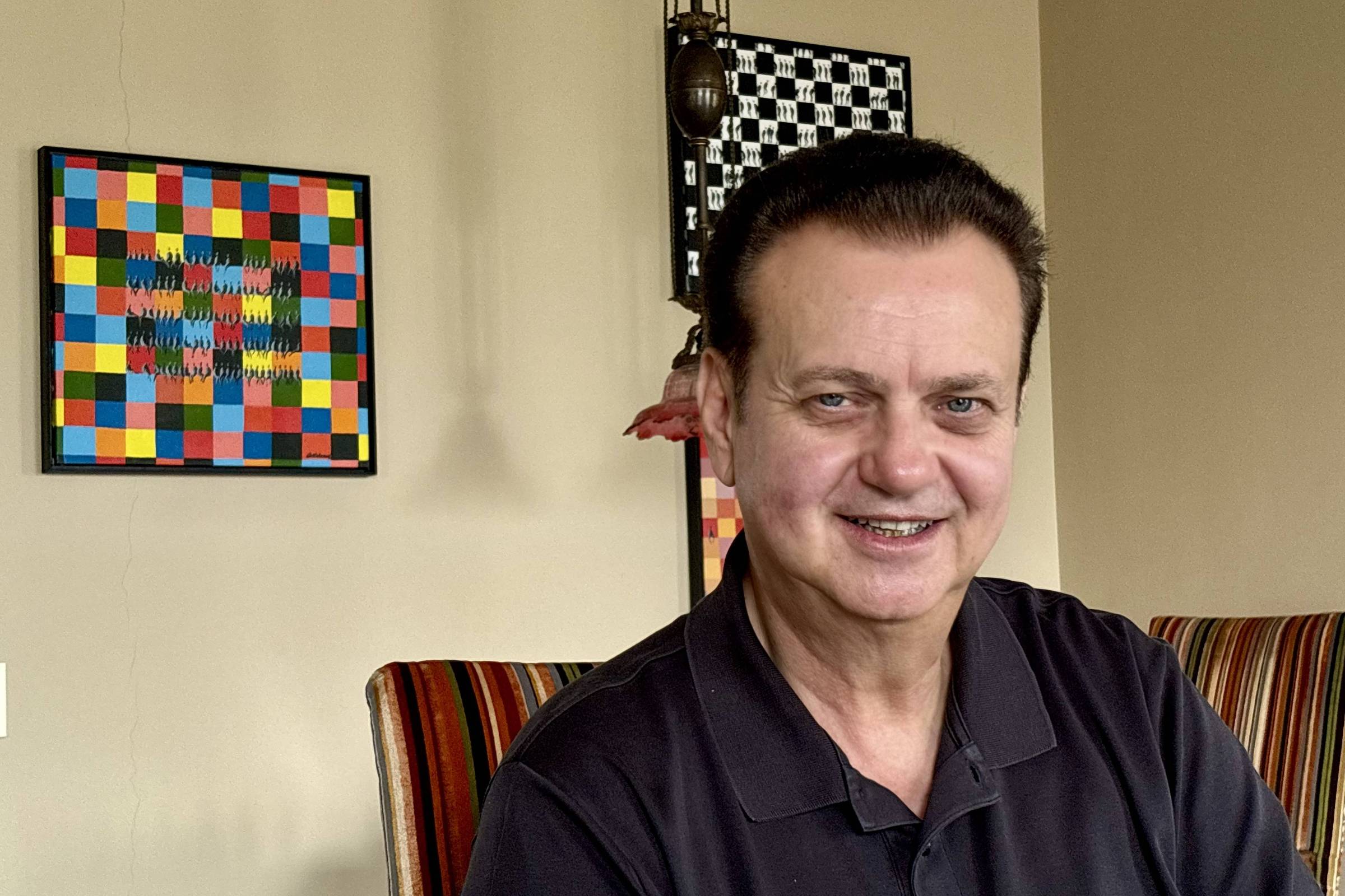 Homem de meia-idade com cabelo escuro penteado para trás, vestindo camisa polo preta, sentado em cadeira com estofado listrado em tons terrosos. Ao fundo, parede bege com dois quadros coloridos, um com padrão quadriculado multicolorido e outro em preto e branco.