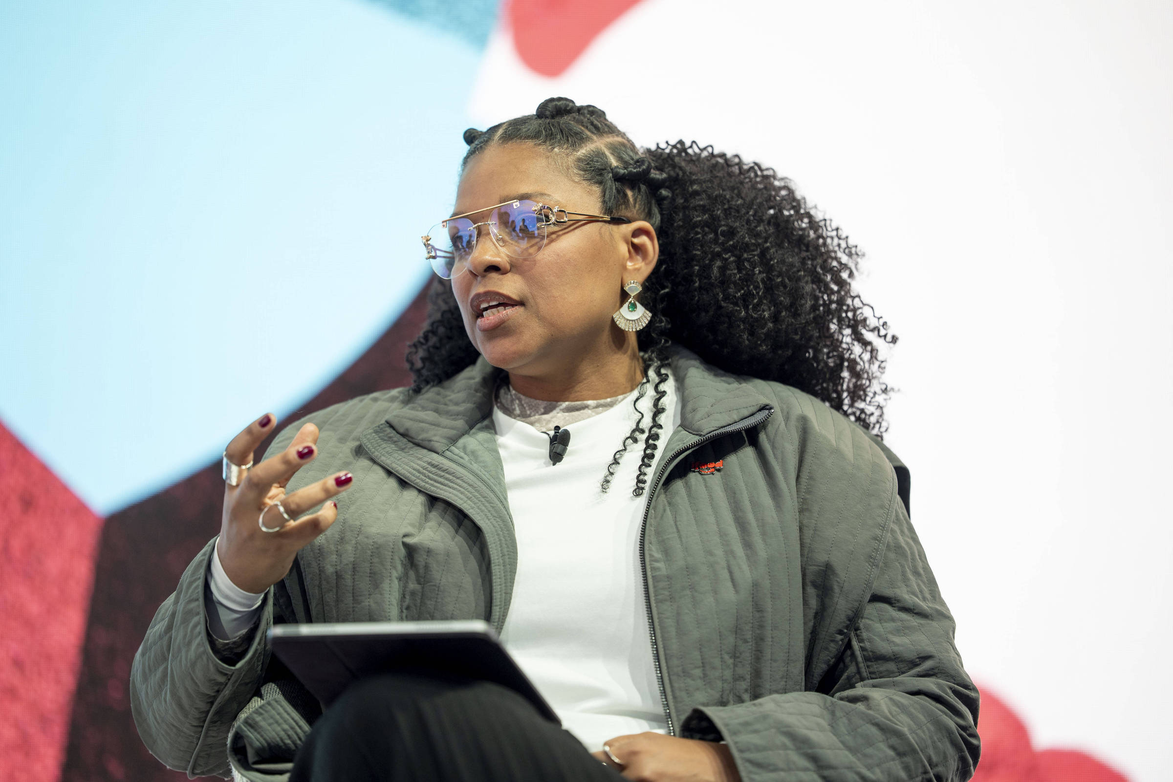 Mulher negra com cabelo cacheado preso parcialmente, usando óculos e brincos, veste jaqueta cinza e camiseta branca, segura tablet no colo e gesticula durante fala em ambiente claro.