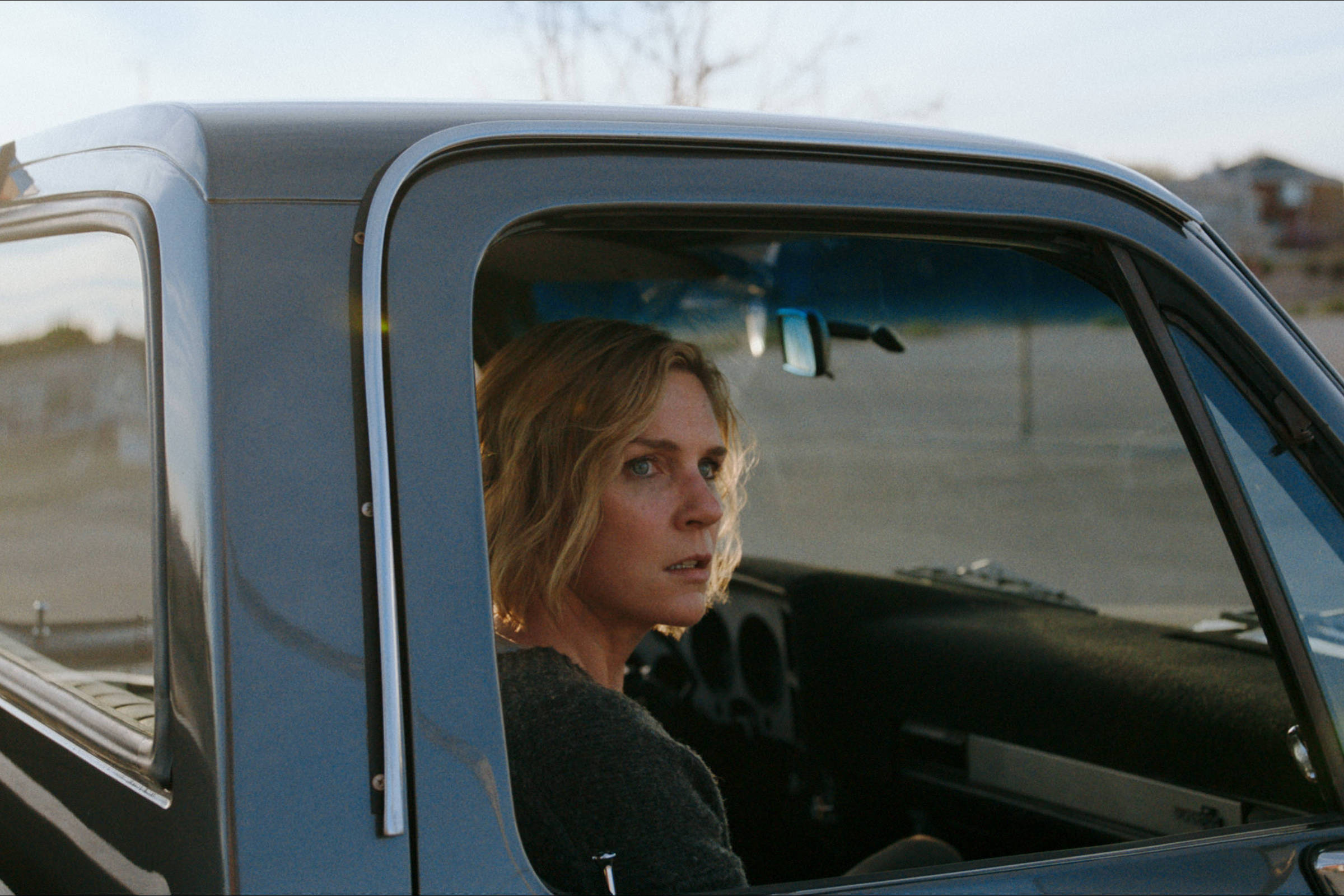 Mulher de cabelos loiros e expressão séria está sentada no banco do motorista de uma caminhonete preta estacionada em área aberta durante o dia. O interior do veículo é visível pela janela aberta, e ao fundo há casas e árvores desfocadas.